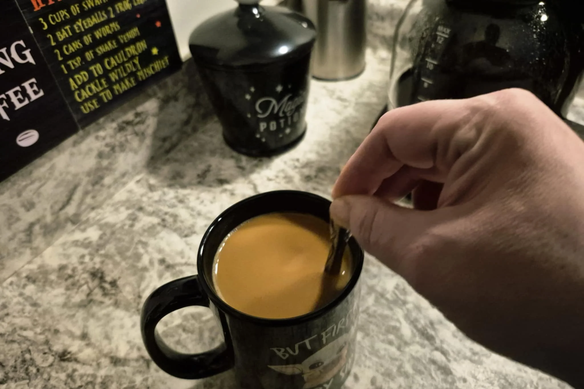 A hand stirring coffee in a black Baby Yoda mug on a kitchen counter, the grinder and drip coffee maker behind it, capturing a small moment of the morning routine.