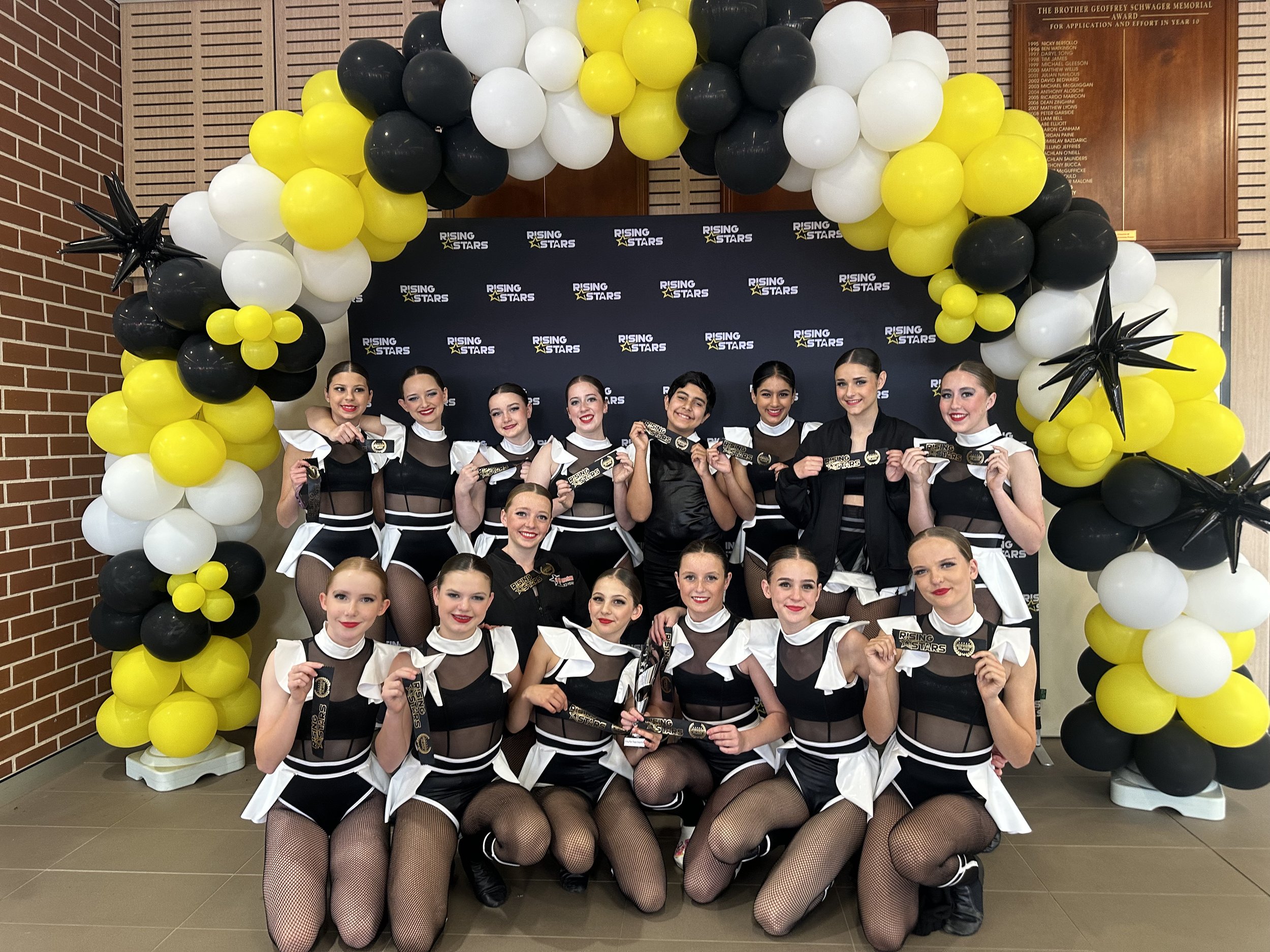 Group of young female dancers in black and white costumes holding medals, posing under black, white, and yellow balloon arch at a dance competition.