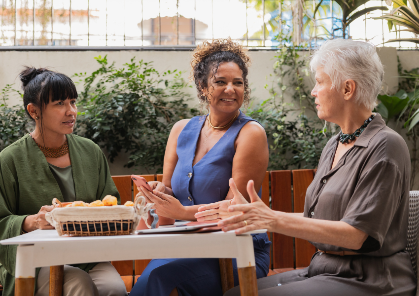 Three women sitting at a white table outdoors, engaged in conversation, with greenery and a window in the background.
