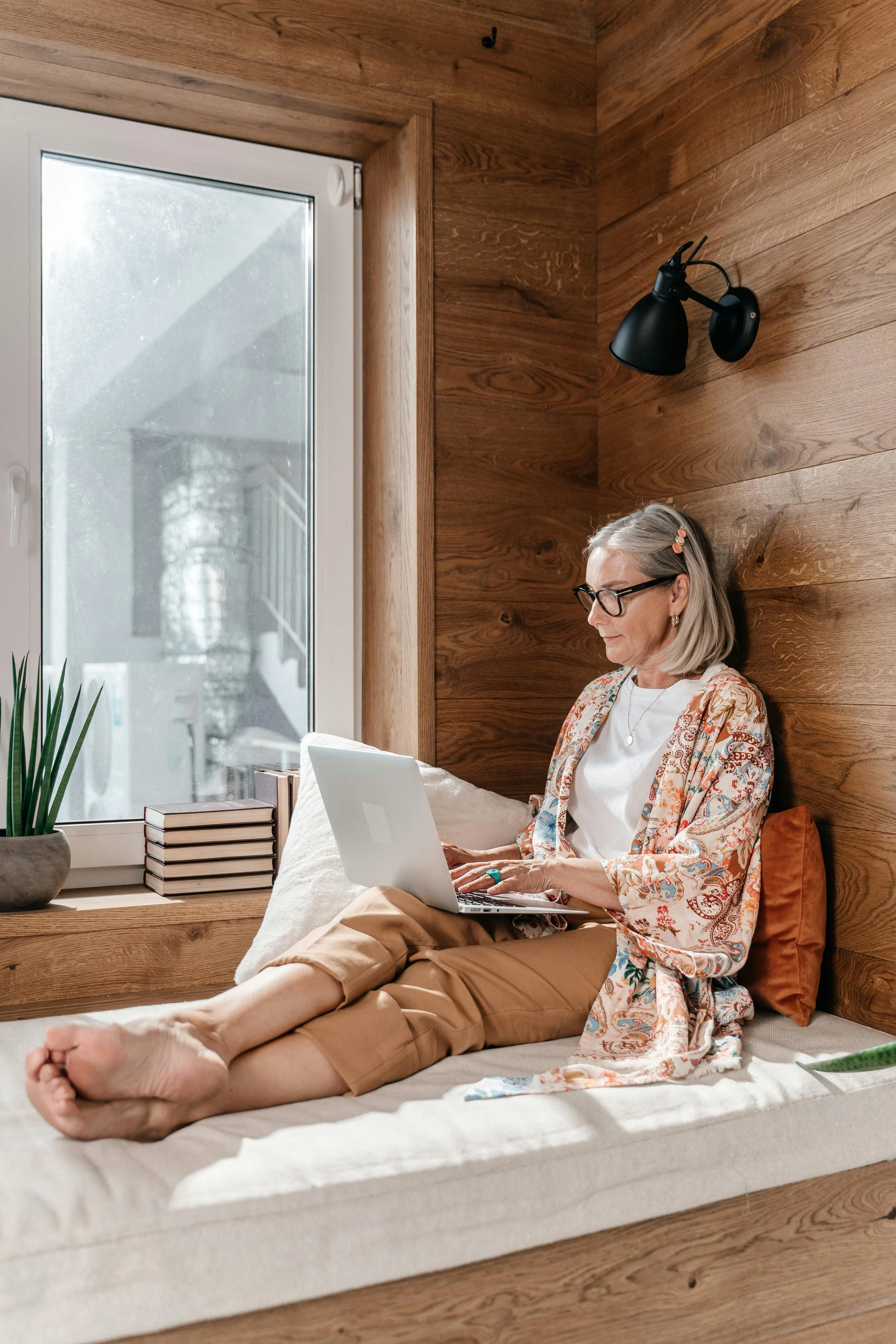 A woman with gray hair, glasses, and a floral-patterned jacket sitting on a bed with her feet stretched out, working on a laptop. She is in a room with wooden walls, a large window, and a black wall-mounted lamp.