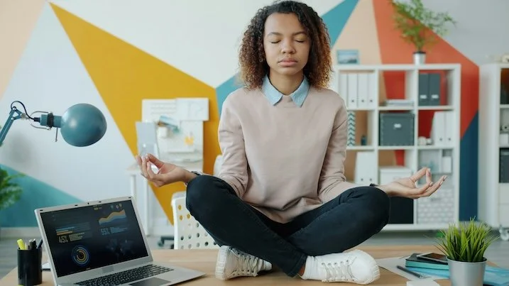 Young woman with curly hair sitting crossed legged on desk in meditation position with eyes closed. Laptop open beside her. Office shelves in background.