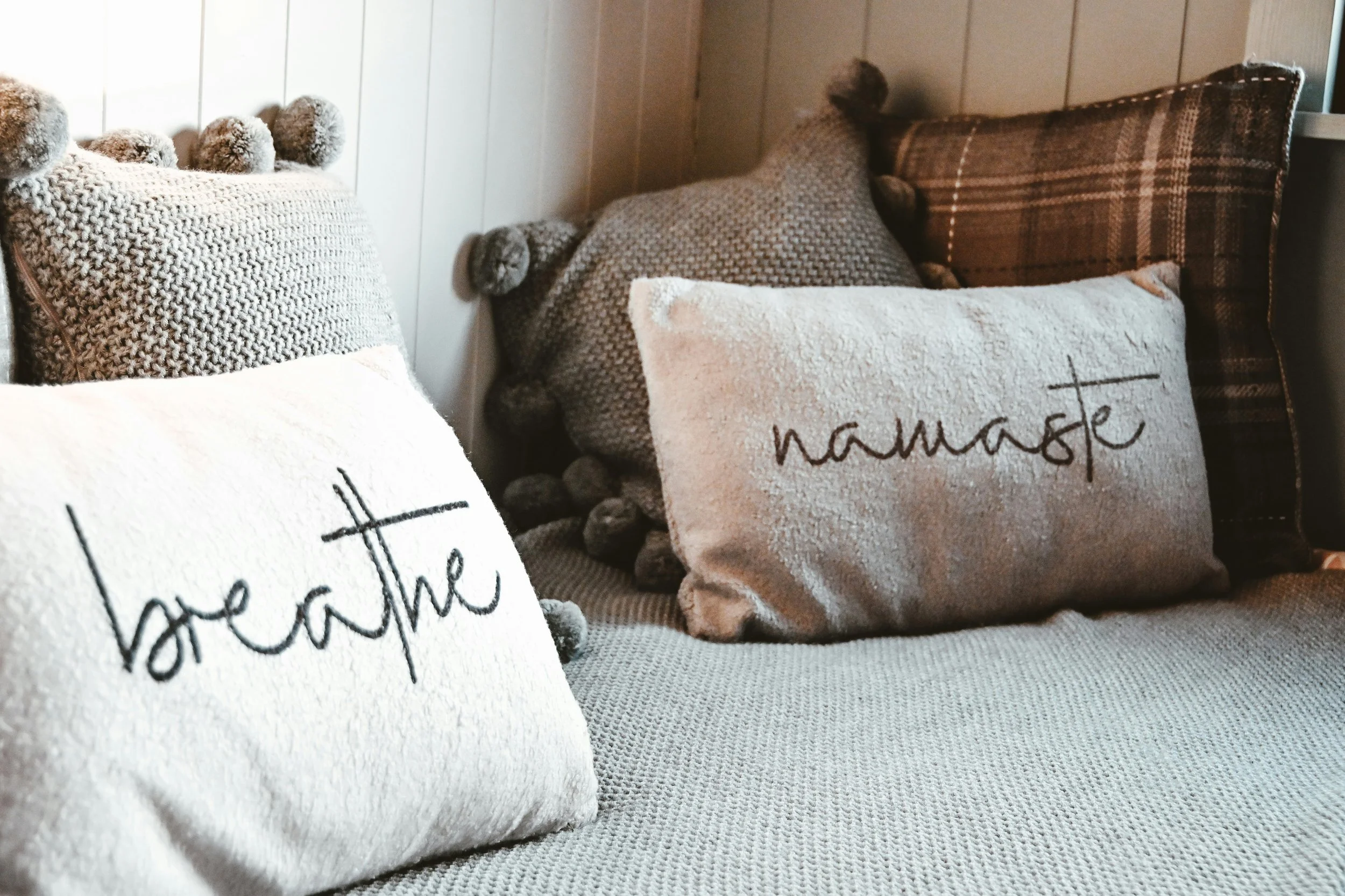 Two cream cushions sitting on a grey blanket. One with word breathe on it, the other, the word namaste. A few brown cushions, two with tassles, one brown tartan, sit behind them.