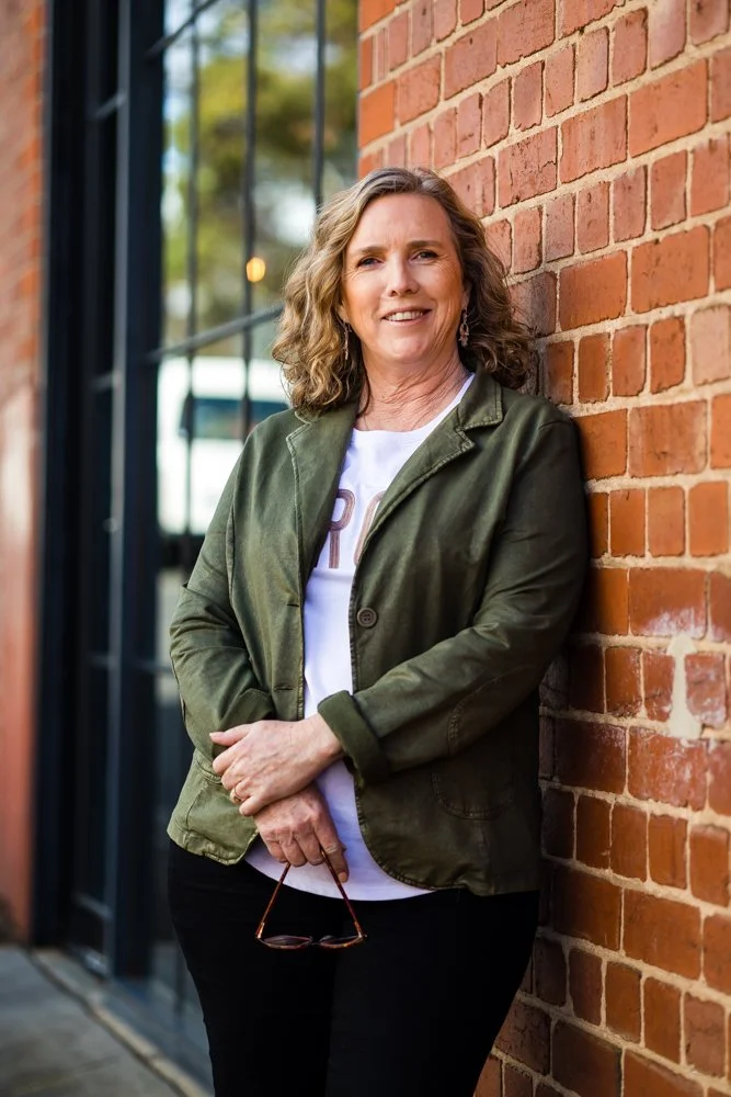 photo of woman in white tee, covered by olive green jacket. She is leaning against a red brick wall with glass window in background. Holding a pair of reading glasses and smiling.