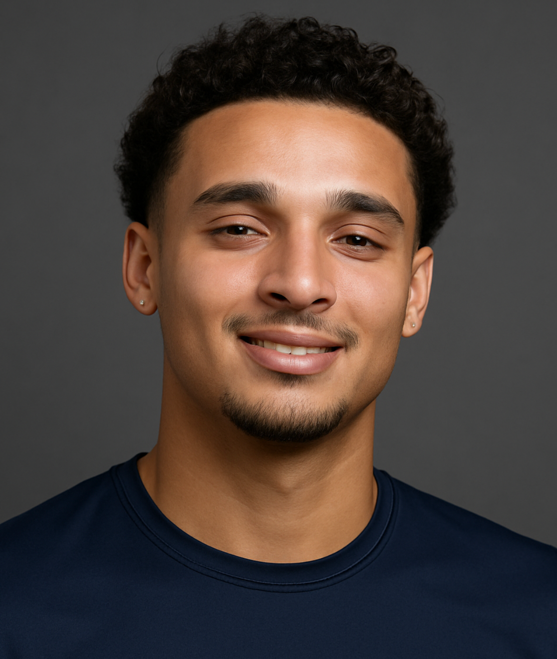 A young man with short curly hair, light brown skin, and small earrings, smiling against a dark gray background. He is wearing a dark blue shirt.