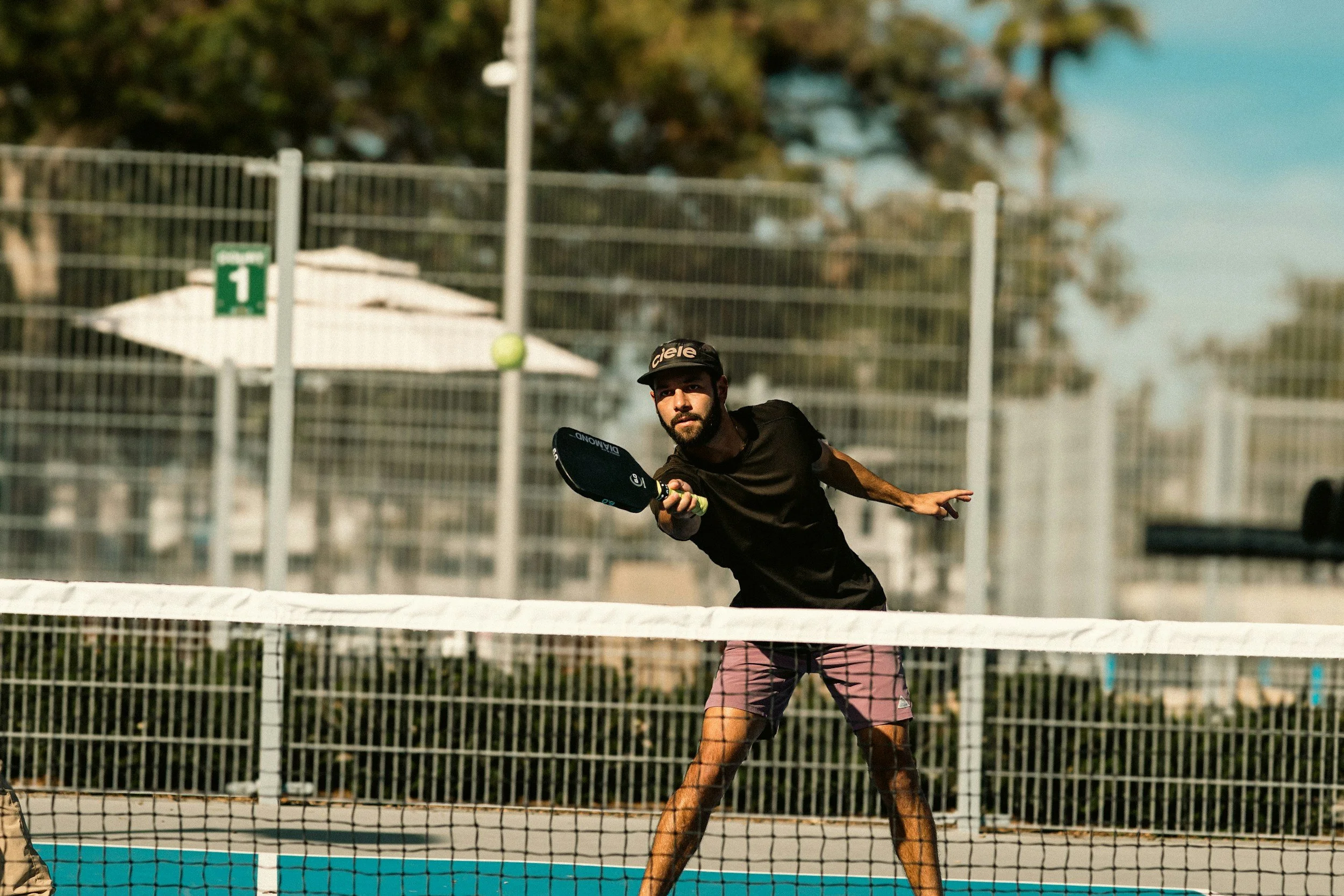 A man playing tennis on an outdoor court with a fence, holding a tennis racket, wearing a cap, black shirt, and patterned shorts, with tennis ball mid-air near his racket.