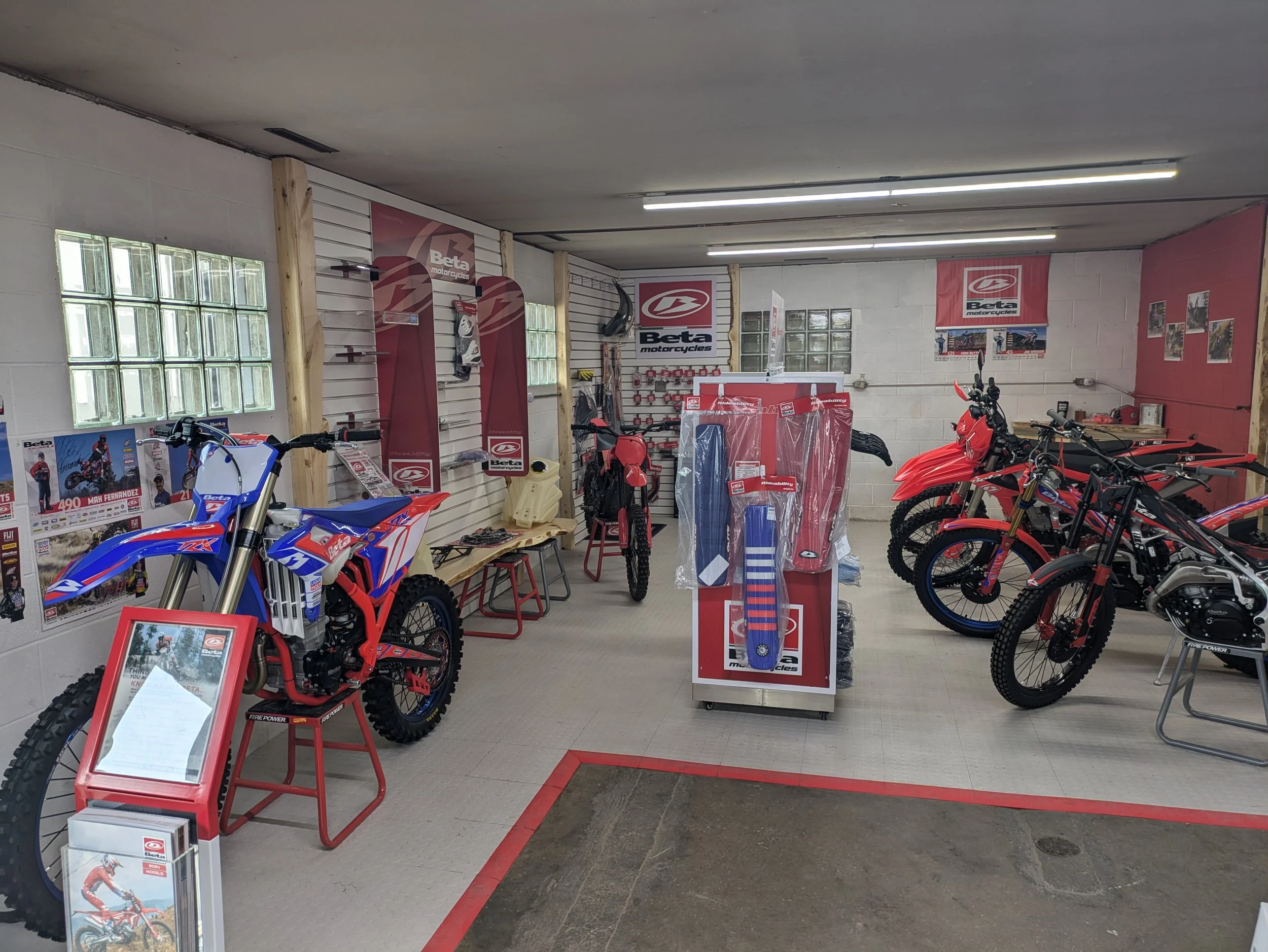 Indoor motorcycle showroom with motocross bikes, accessories, and promotional posters, featuring a blue and red motocross bike, and multiple red dirt bikes.