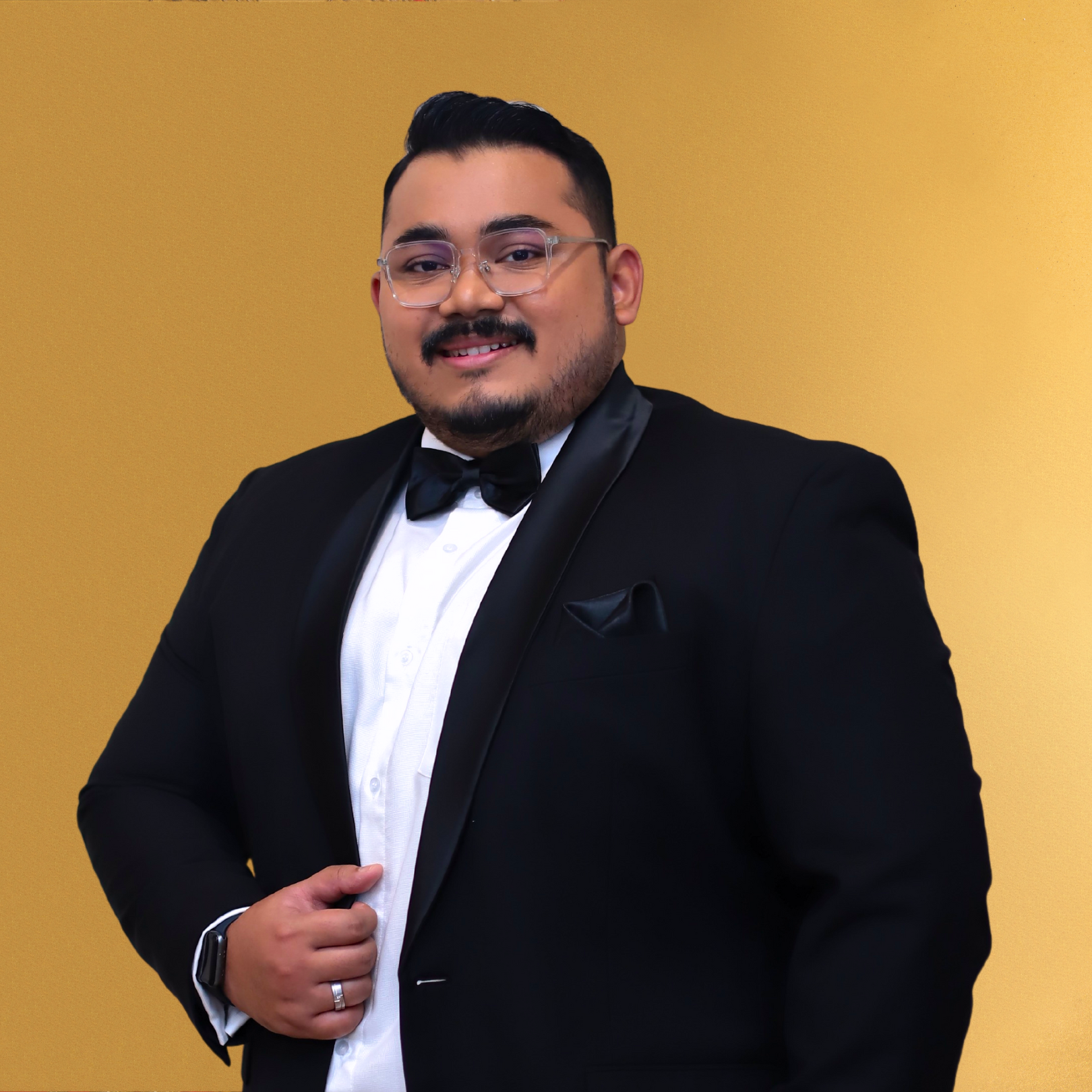 A man wearing a black tuxedo with a white shirt and black bow tie, posing confidently against a yellow background.
