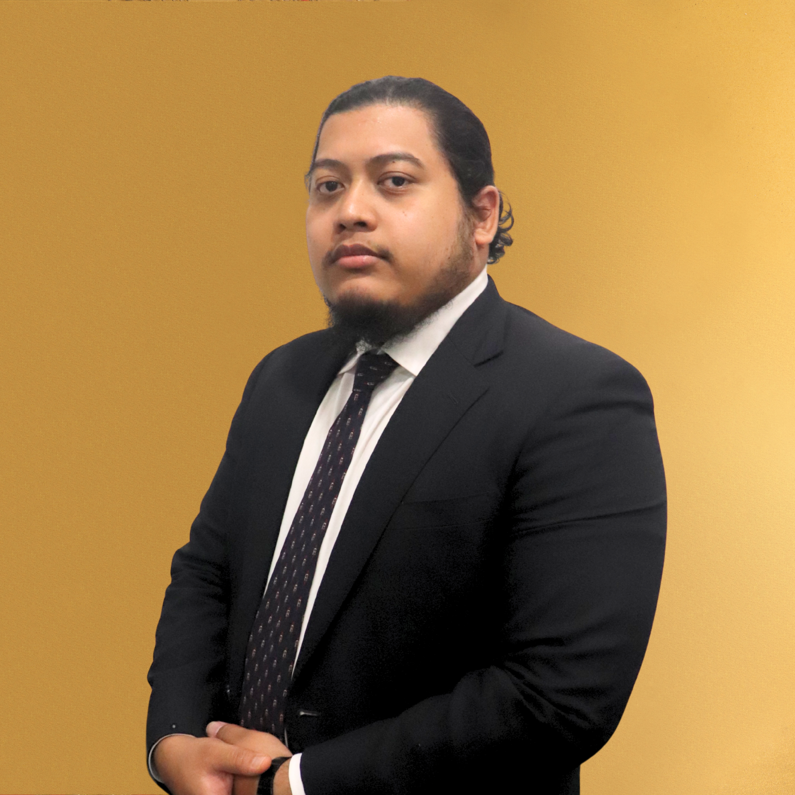 A man in a black suit, white shirt, and patterned tie standing against a yellow background.