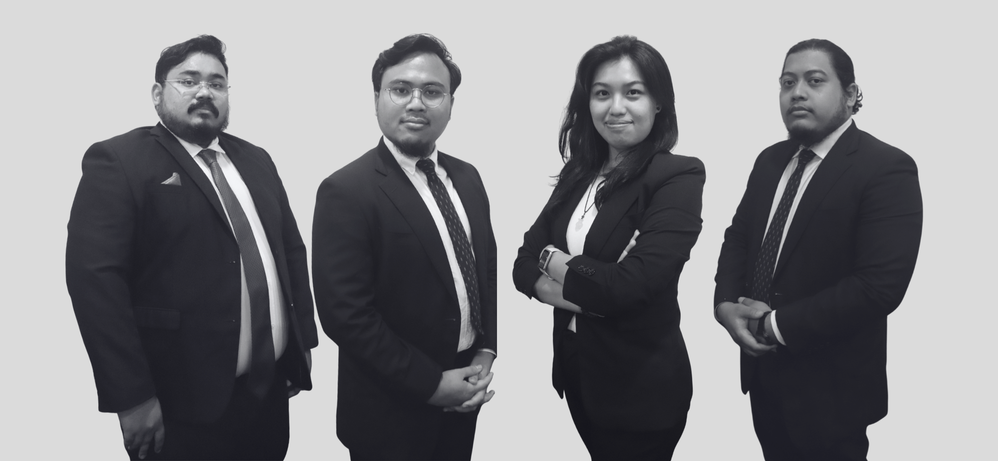 Four professionals dressed in formal business suits standing against a plain light grey background.