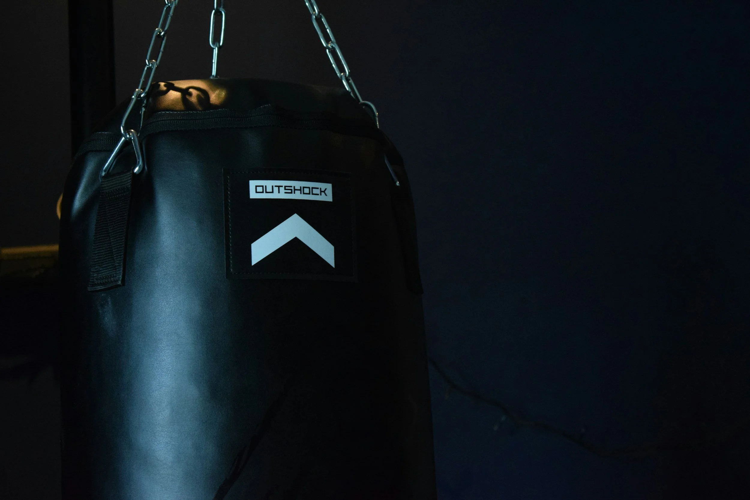 Black punching bag with a white logo reading 'OUTSHOCK' and a black arrow symbol, hanging from metal chains against a dark background.