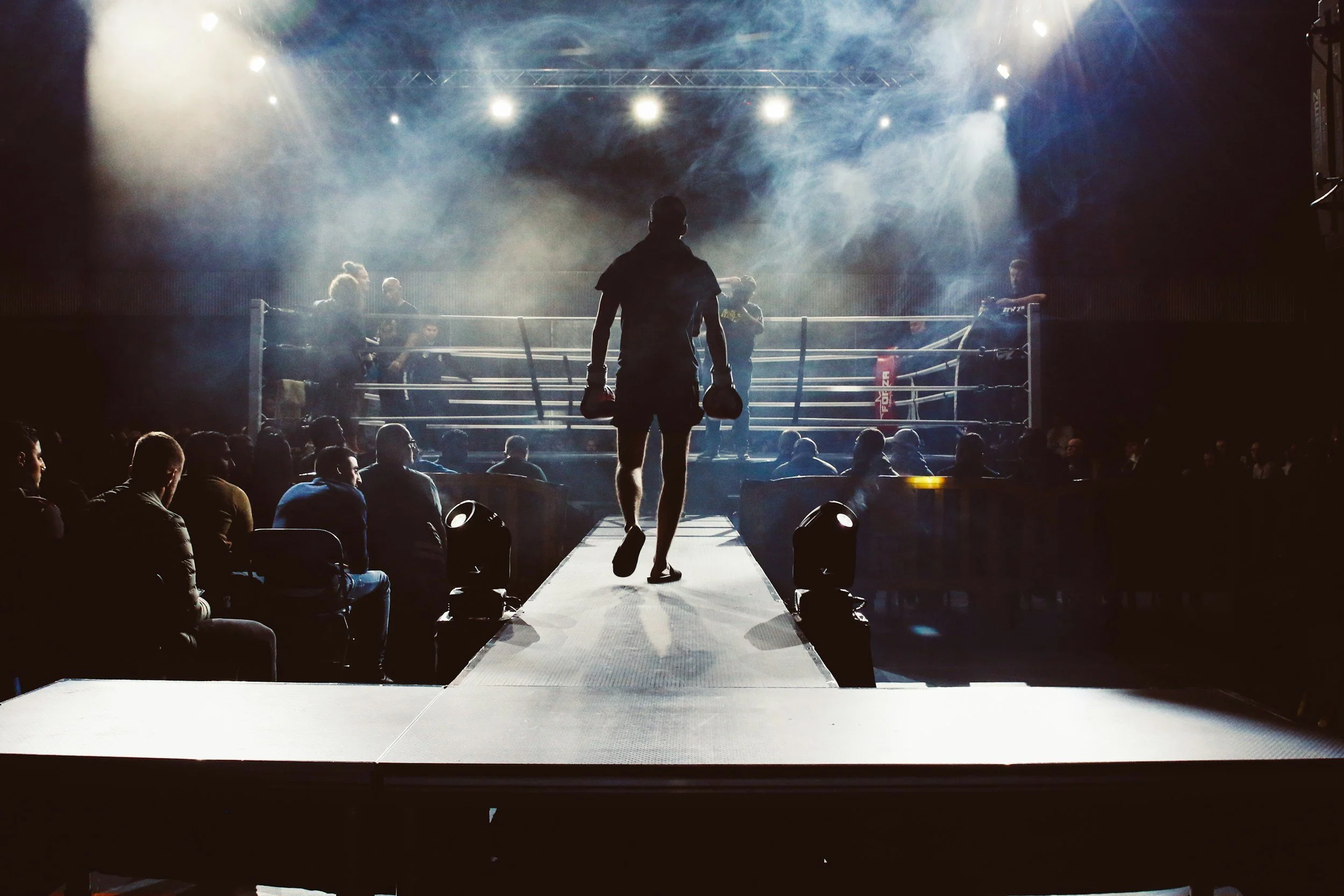 A boxer holding gloves walks onto the boxing ring as the audience watches, with spotlights and smoke effects in the background.