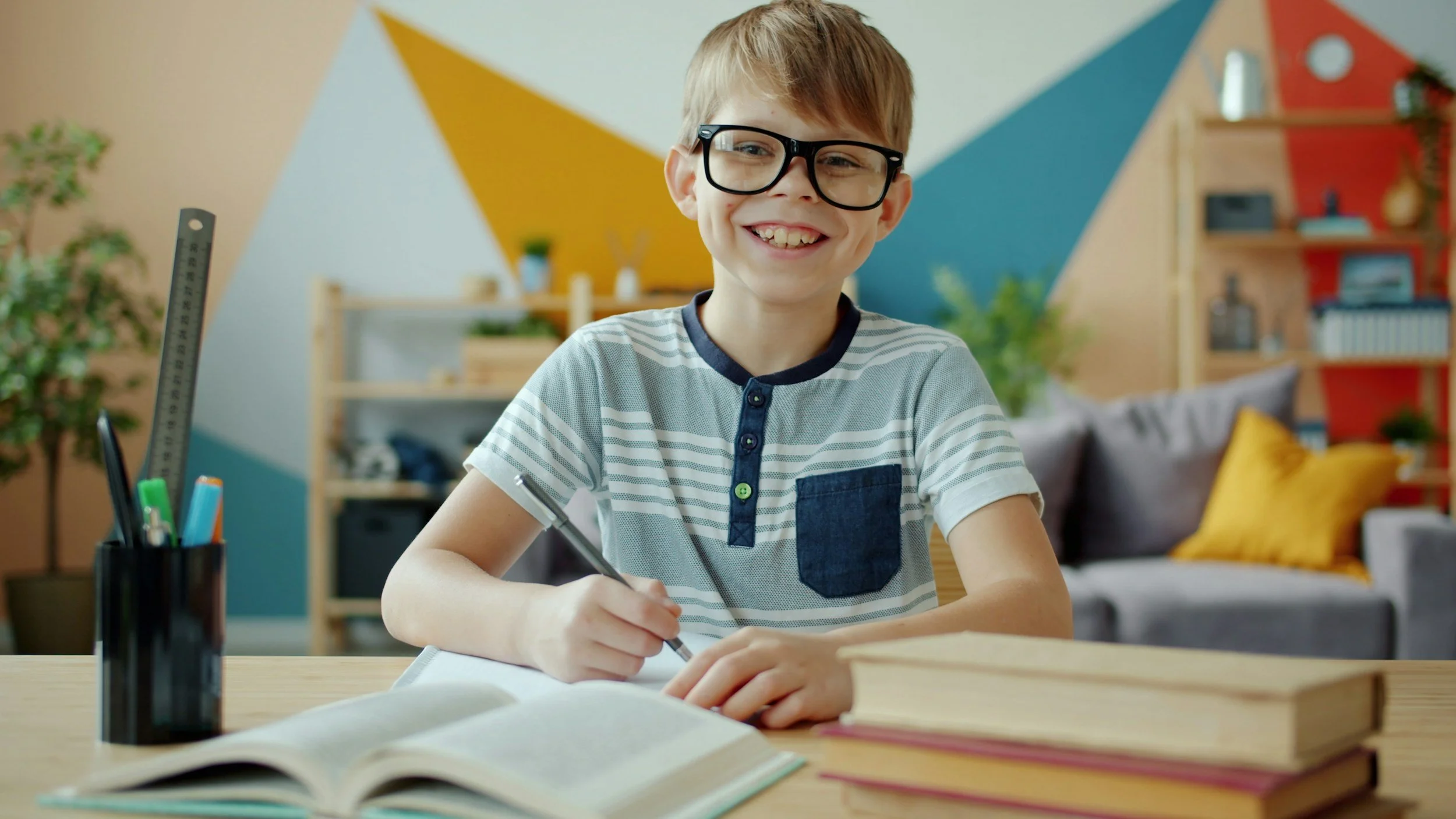 A smiling boy with glasses sitting at a desk with books, writing in a notebook. The background is a colorful, modern room with a plant, bookshelf, and decorative pillows.