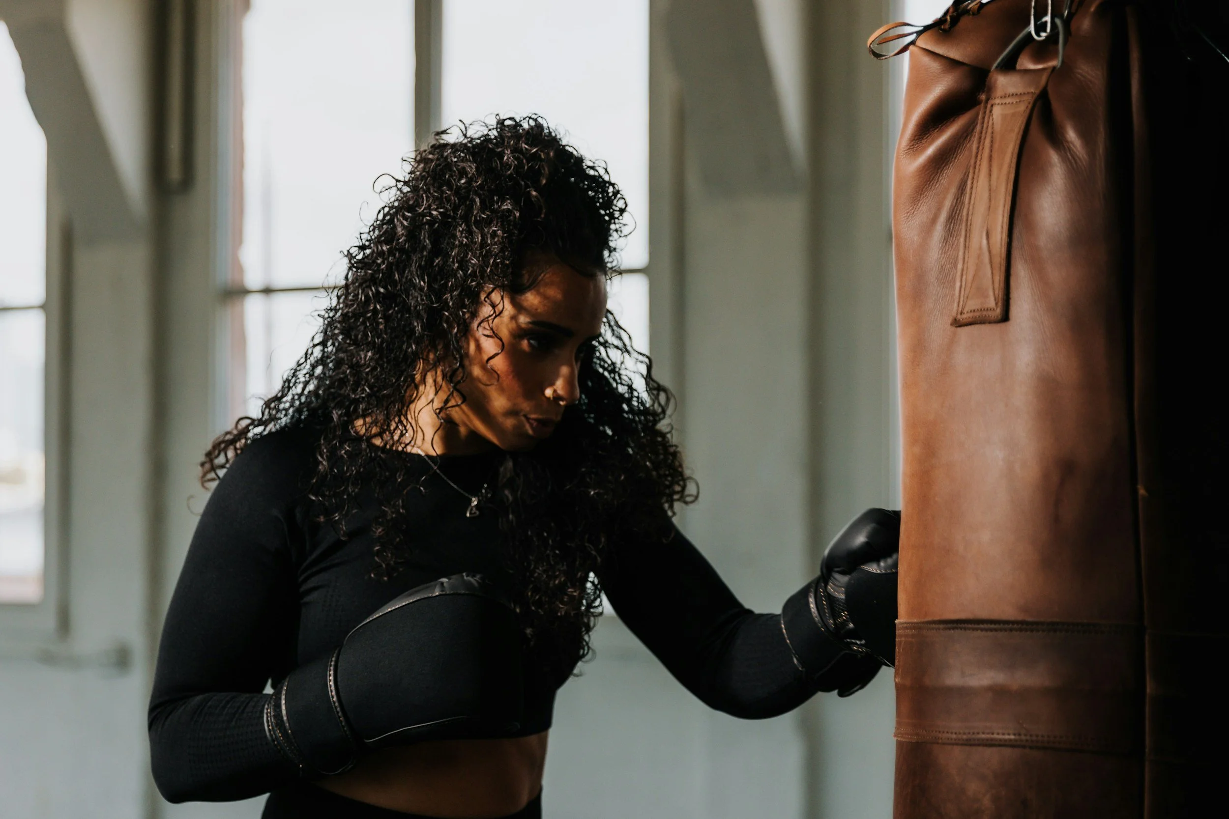 A woman with curly dark hair wearing black athletic clothing and boxing gloves, practicing punches on a brown leather punching bag in a gym with large windows.