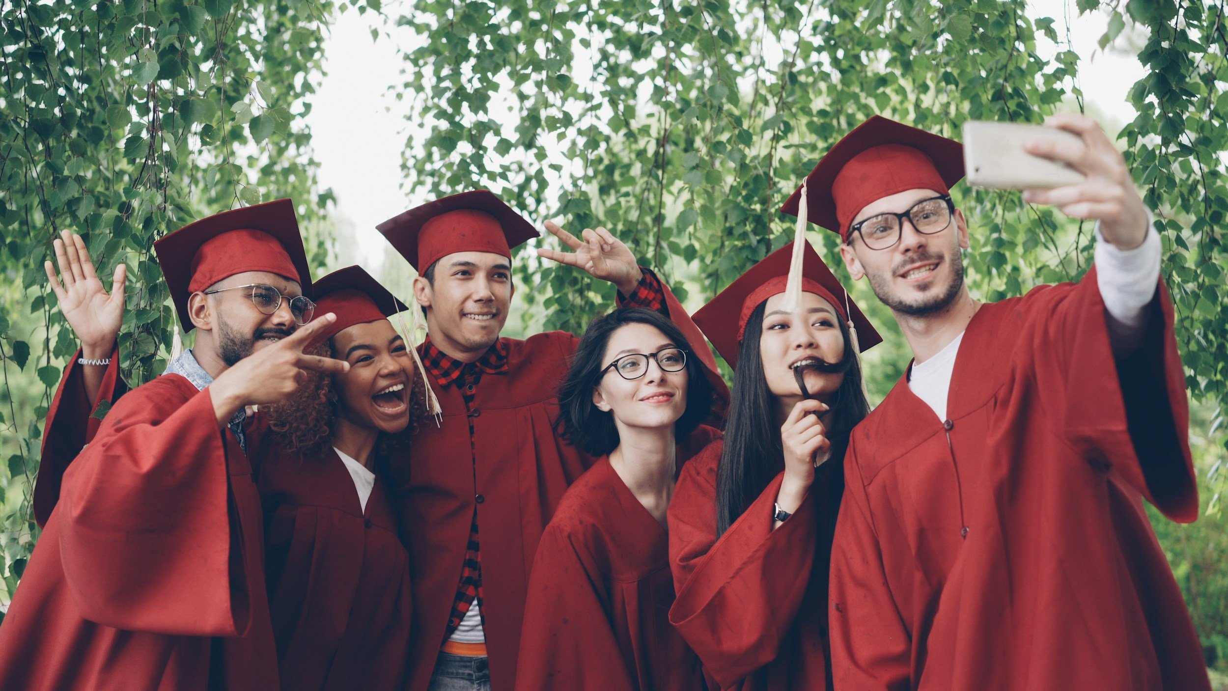 Group of diverse students in red graduation gowns and caps taking a selfie outdoors under green trees.