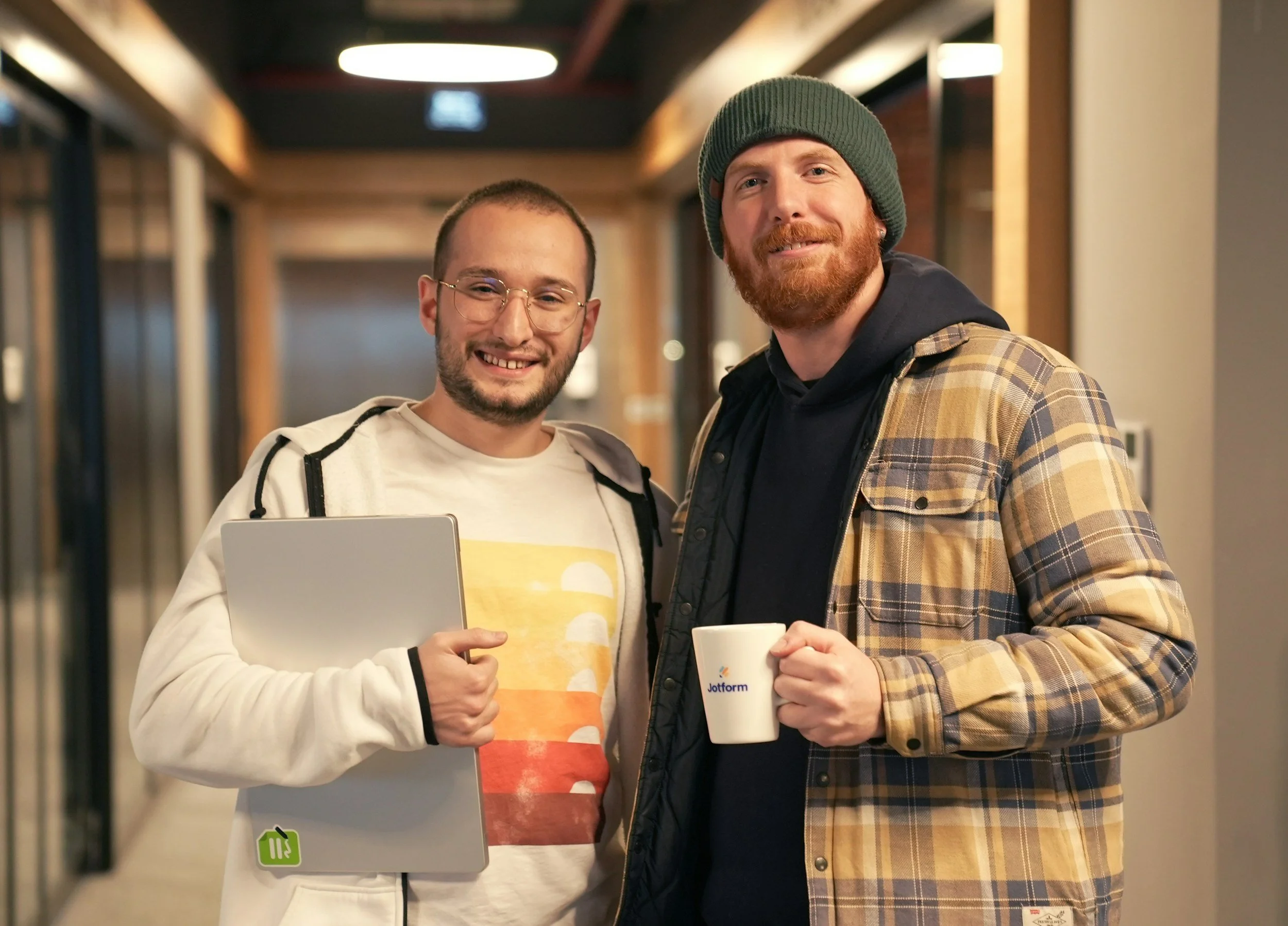Two smiling men standing together in an office hallway, one holding a tablet and the other holding a coffee mug, both casually dressed.