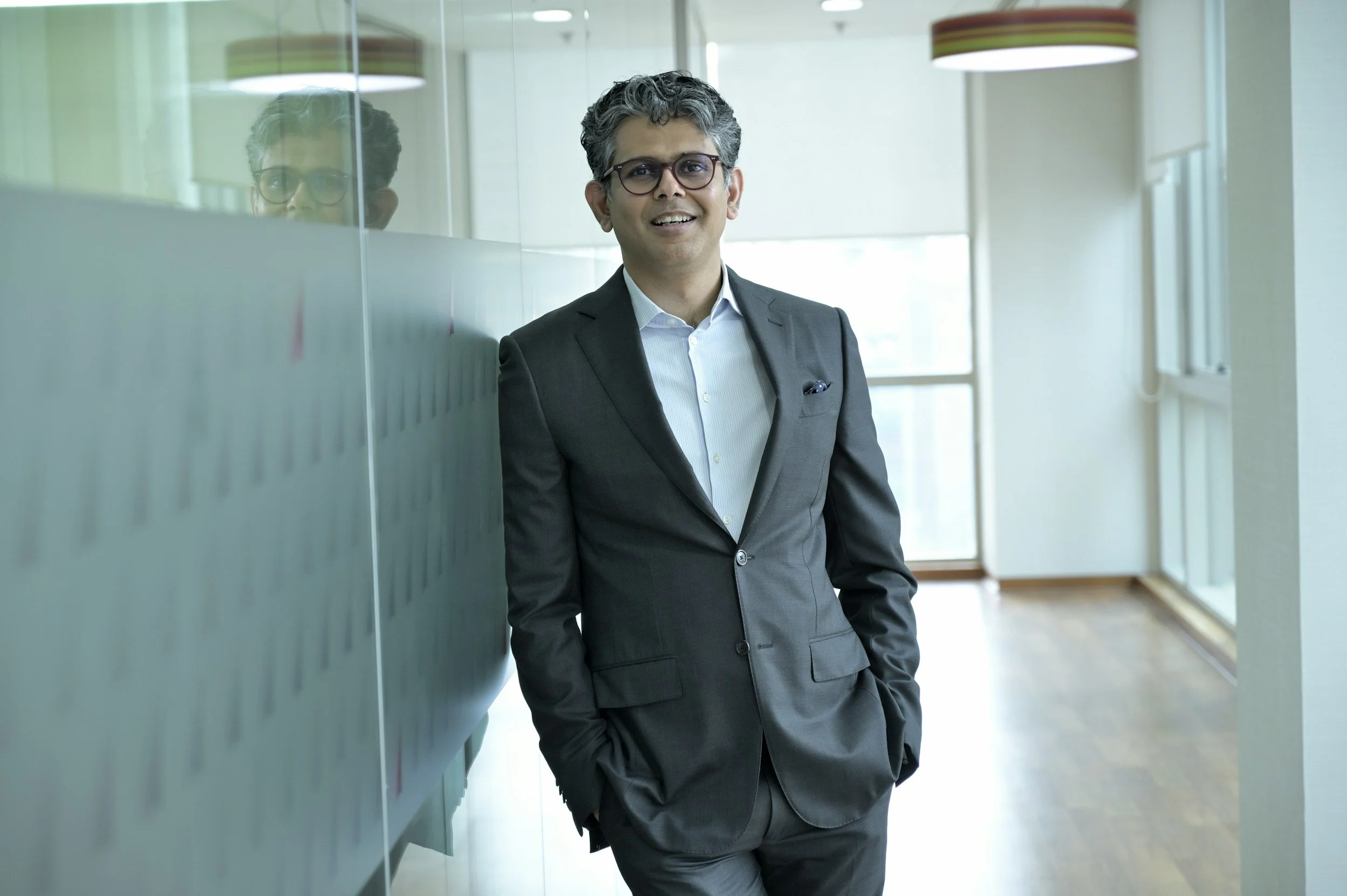 A man in a gray business suit and glasses standing in a modern office hallway, smiling with his hands in his pockets, next to a glass wall.