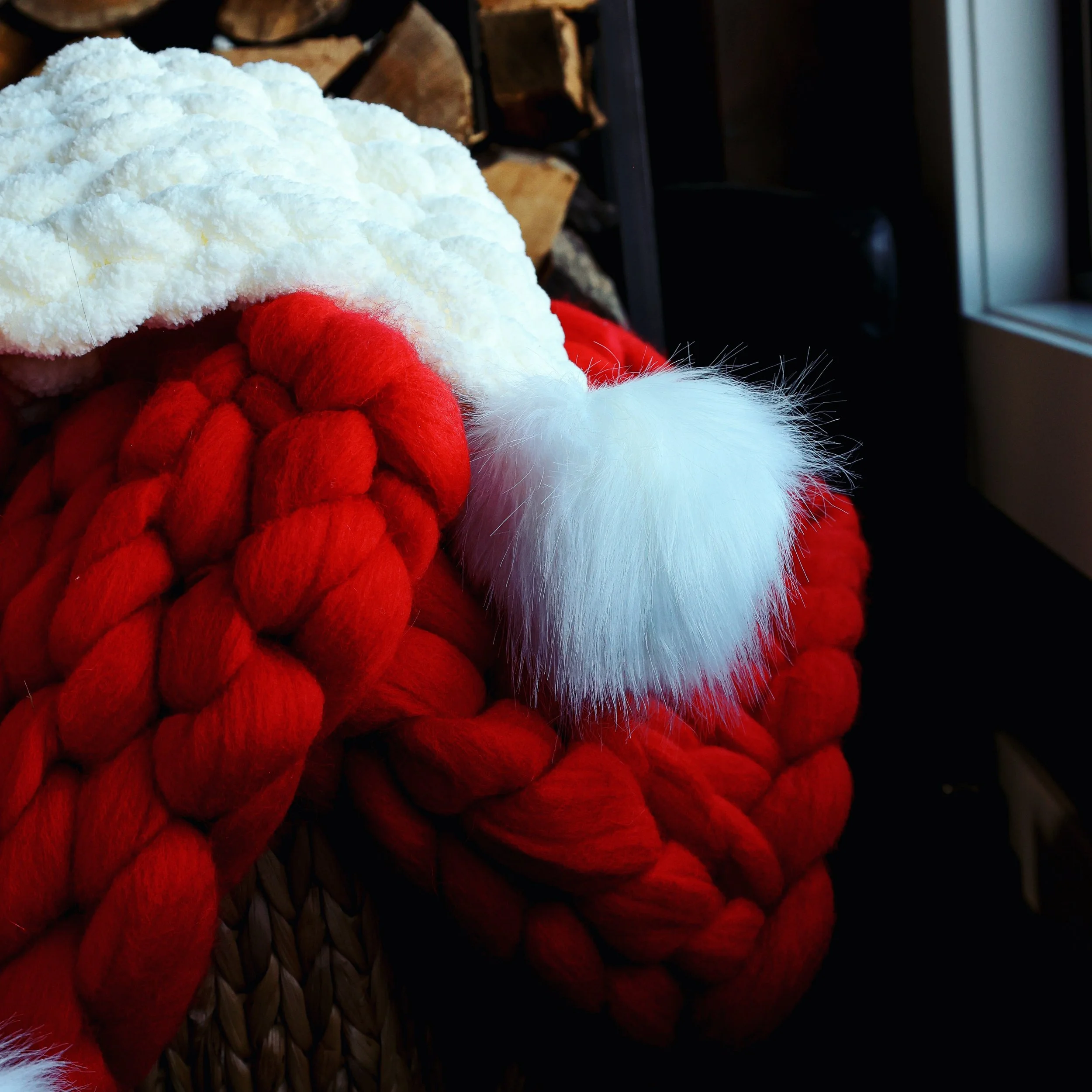 Close-up of Santa hat with red and white fur ball, partially covering a knitted red Christmas stocking. In the background, a basket of firewood and a window are visible.