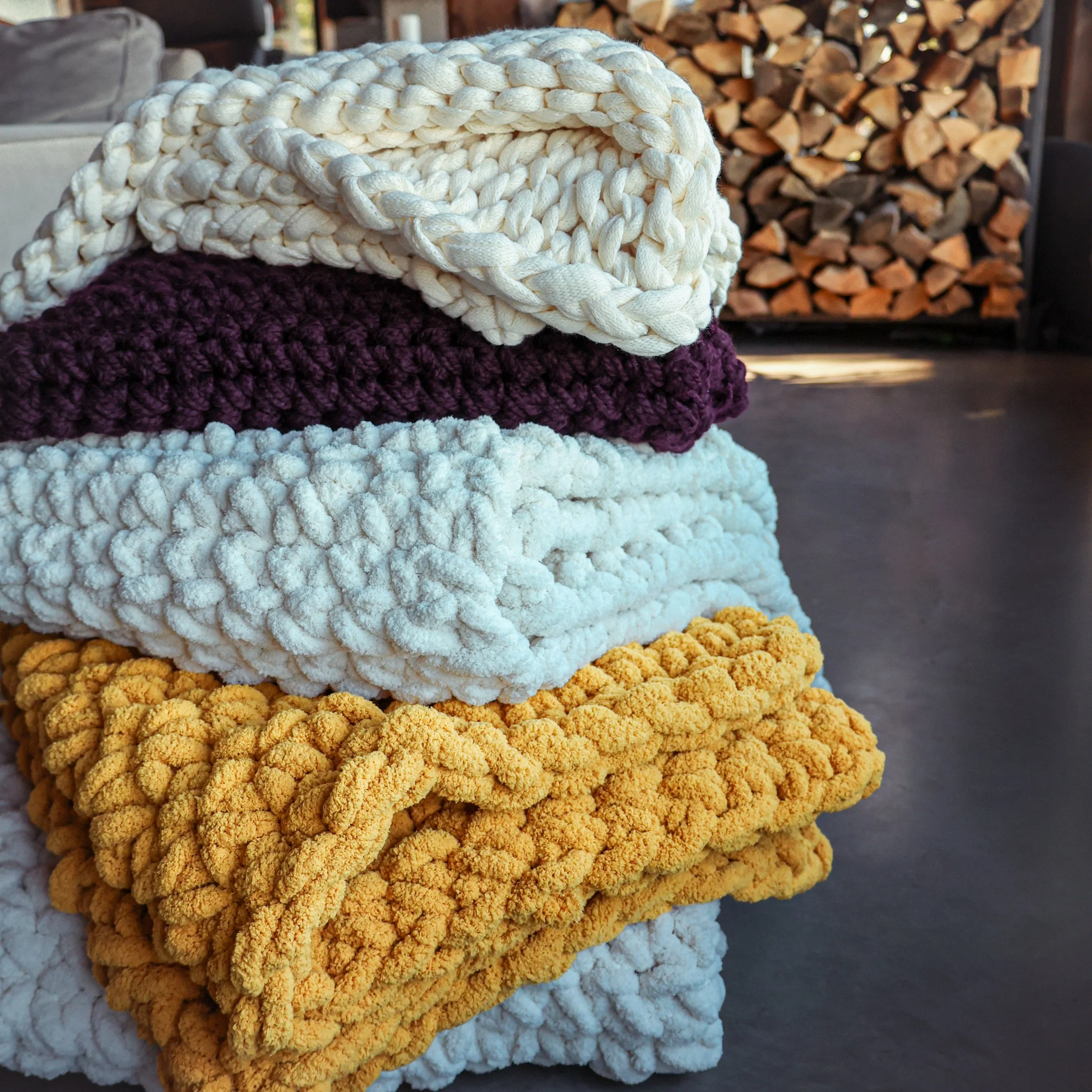 Stacked knitted blankets in cream, purple, white, and yellow colors with a woodpile in the background.