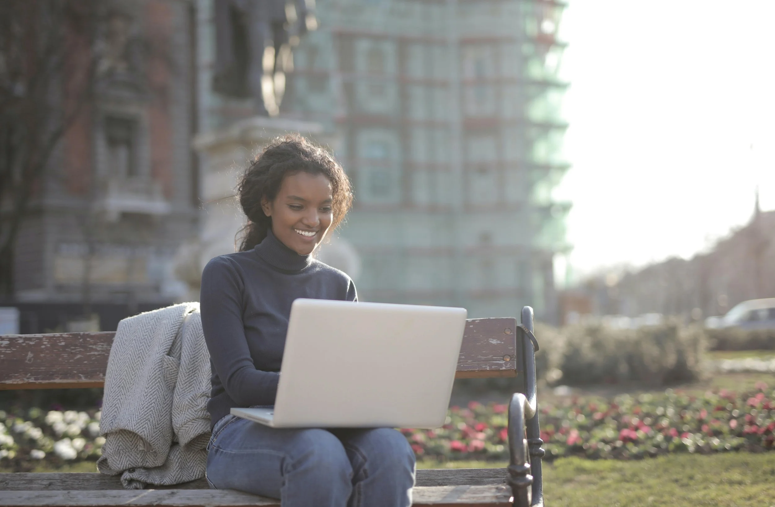 A young woman sitting on a park bench using a laptop, with a jacket draped on the bench beside her, in a city park, smiling at her screen on a sunny day.