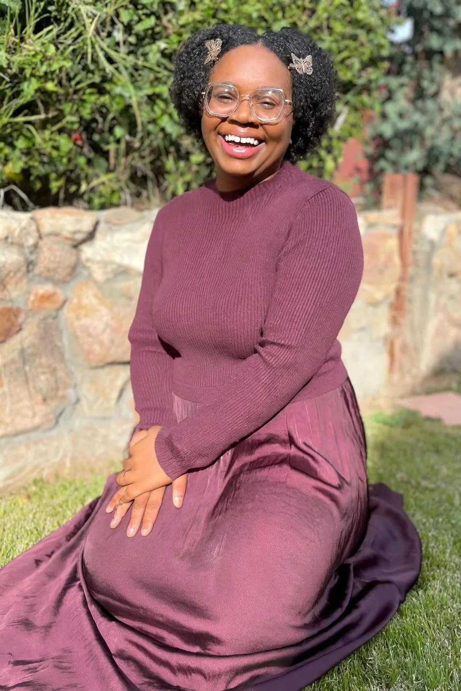 A woman with curly hair and glasses, smiling and sitting on the grass outdoors, wearing a maroon sweater and a matching skirt, with a butterfly hair clip in her hair, in front of a stone wall and green foliage.