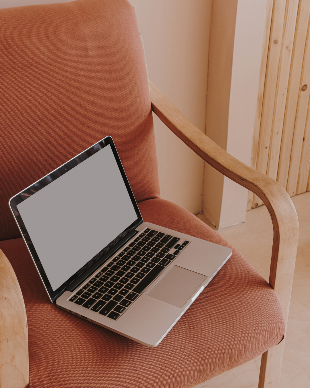 A MacBook laptop with a blank screen resting on a pink upholstered armchair with wooden armrests.