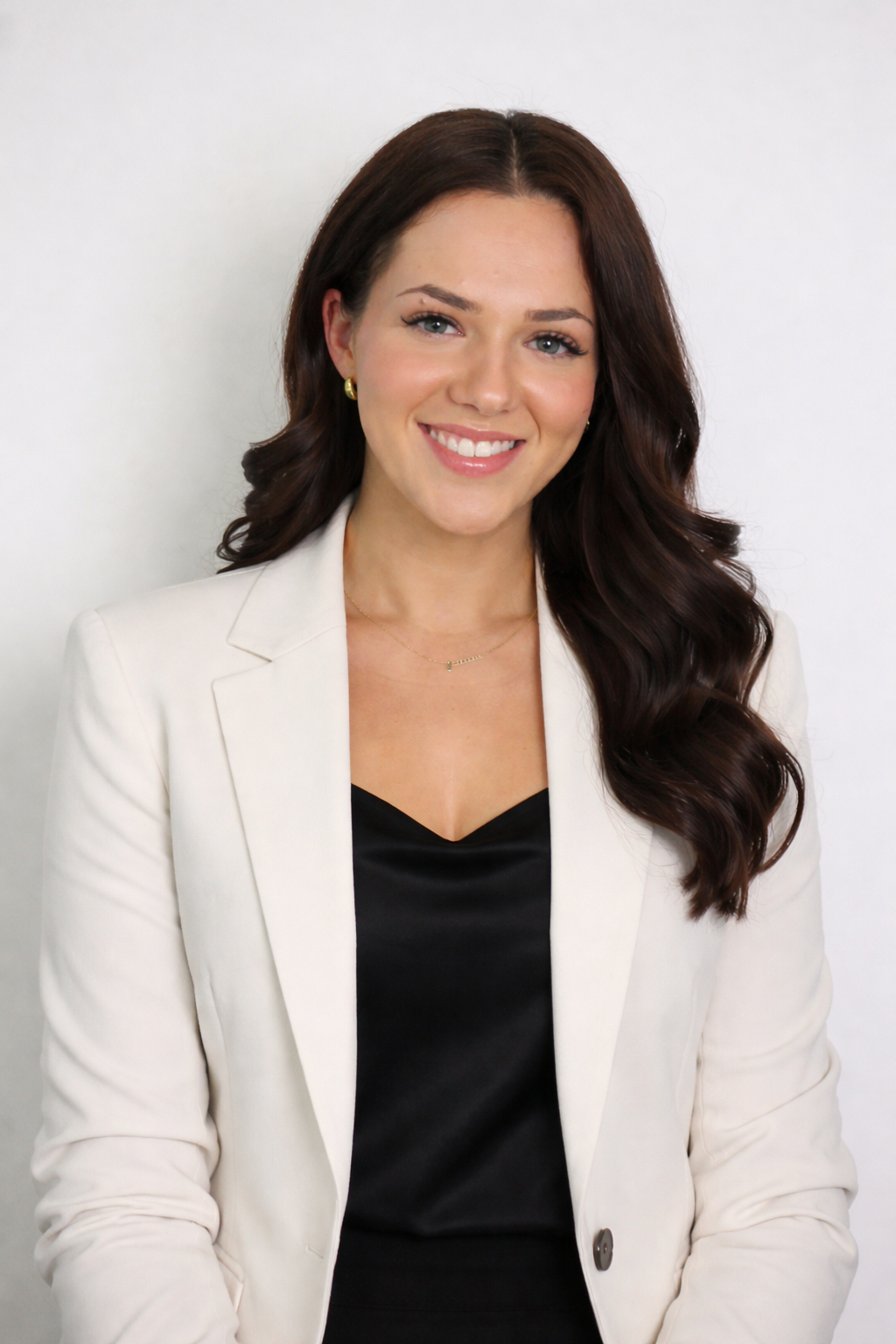 A smiling woman with long brown wavy hair, wearing a white blazer over a black top, gold earrings, a delicate gold necklace, and sitting against a plain white background.