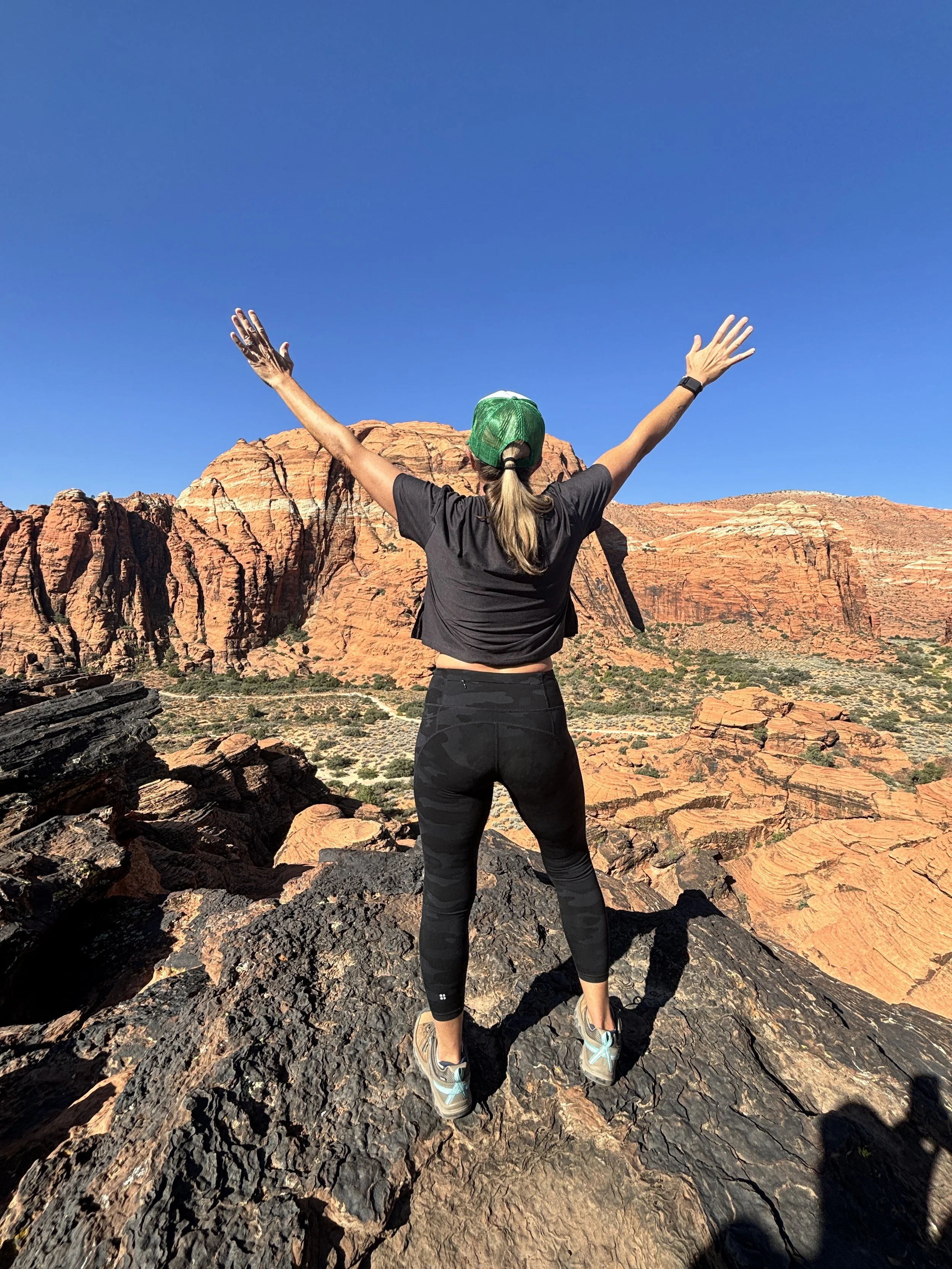 A woman standing on dark volcanic rocks with arms raised, facing away from the camera, overlooking red rock formations and a clear blue sky in a desert landscape.