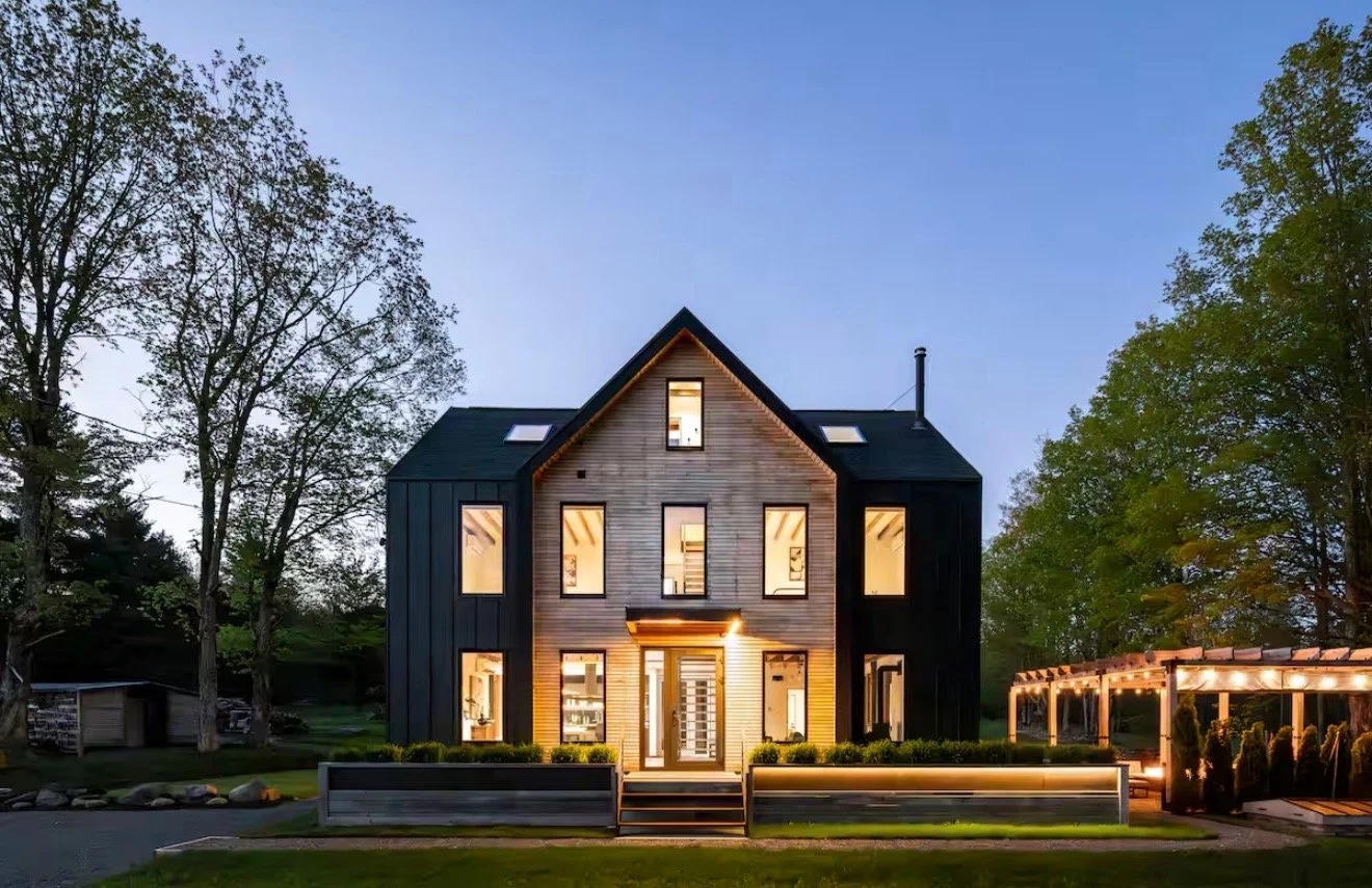 Modern house with large windows and a lit entrance, surrounded by trees and a landscaped yard in evening light.