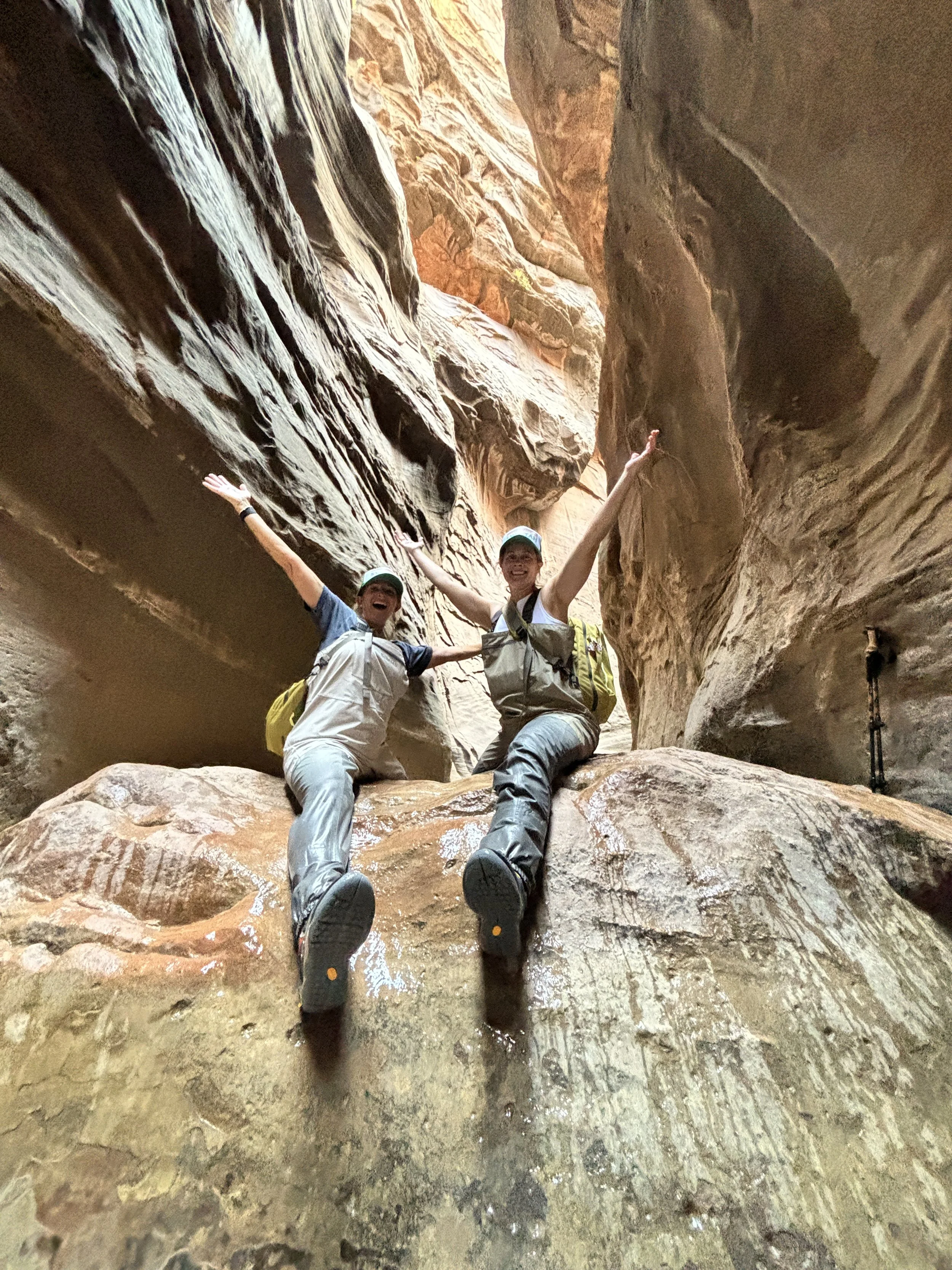 Two women in hiking gear smiling and jumping inside a narrow canyon with smooth, reddish-brown rock walls.