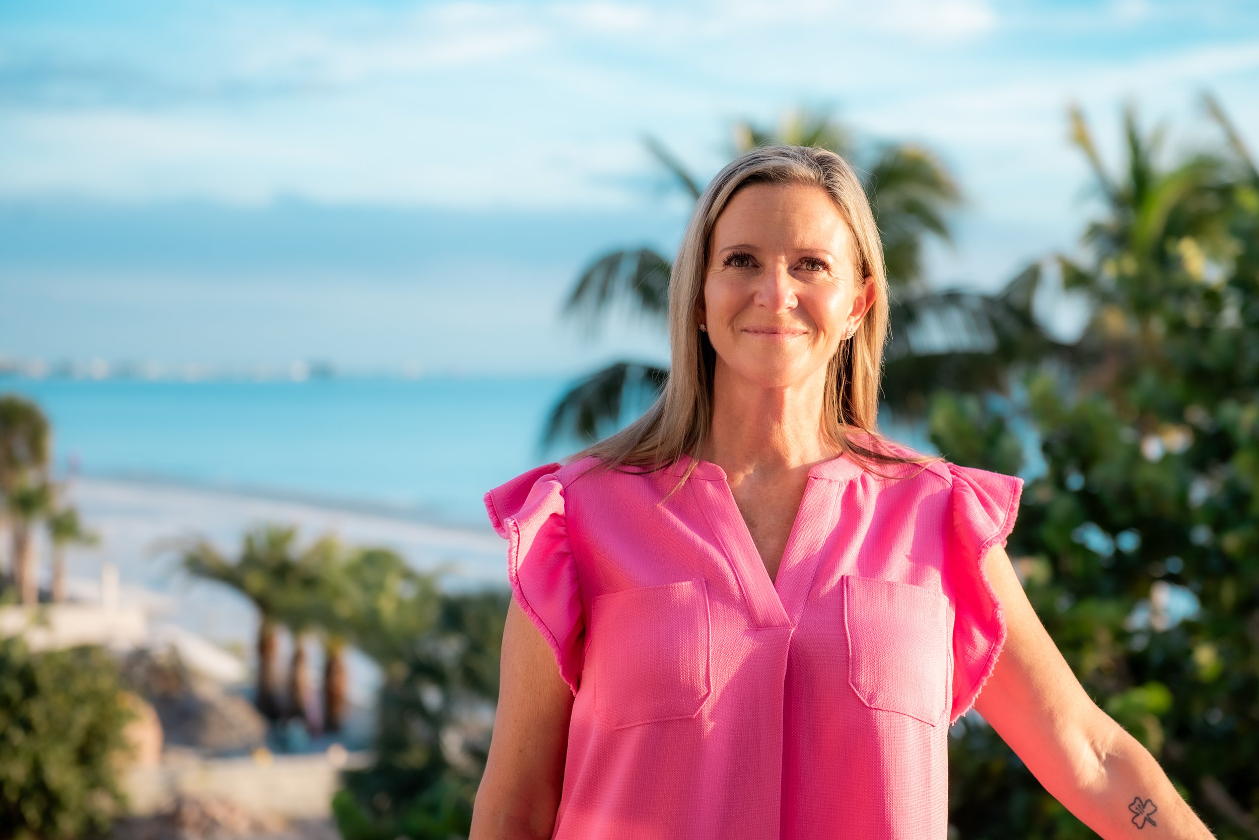 Monica Leonard, a midlife coach, smiling outdoors in a pink top with palm trees and the ocean in the background.