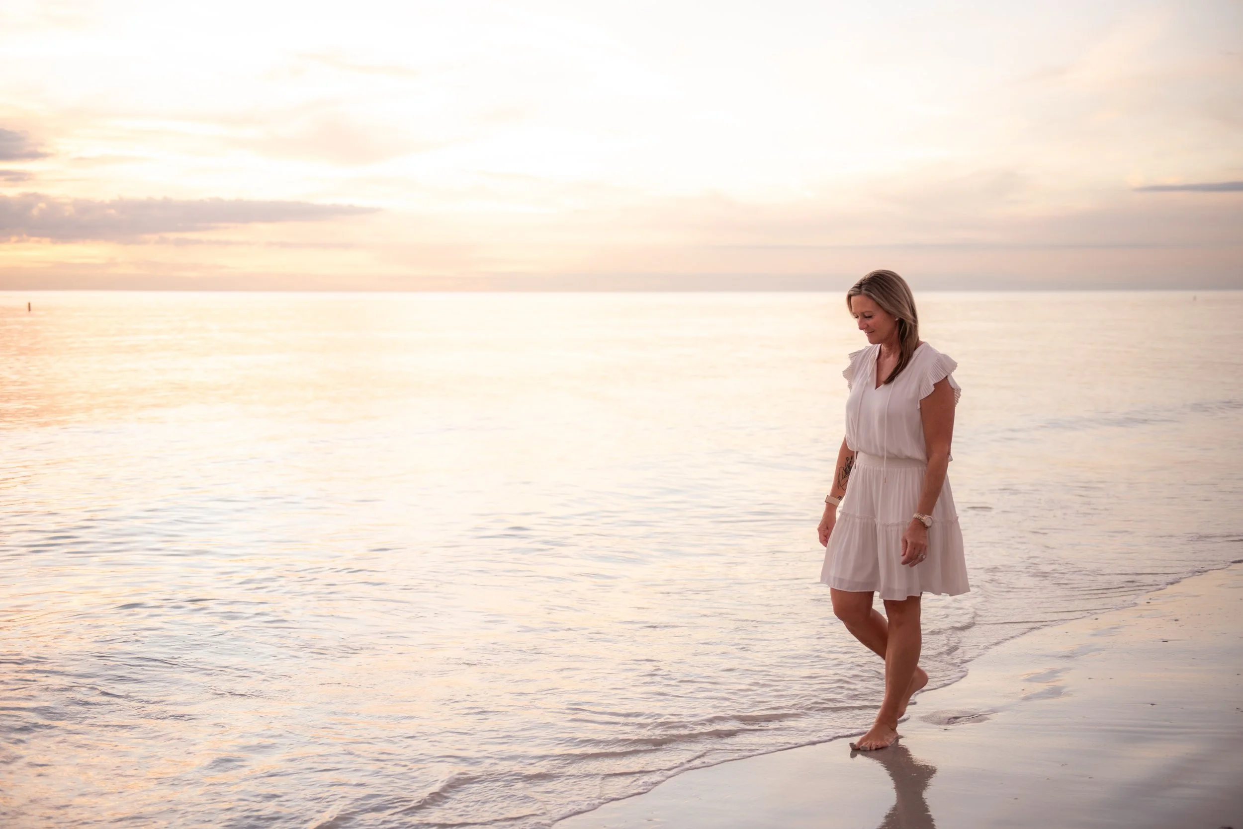Monica Leonard walks barefoot along the shoreline in a white dress at sunset. The ocean is calm with soft golden light across the water.