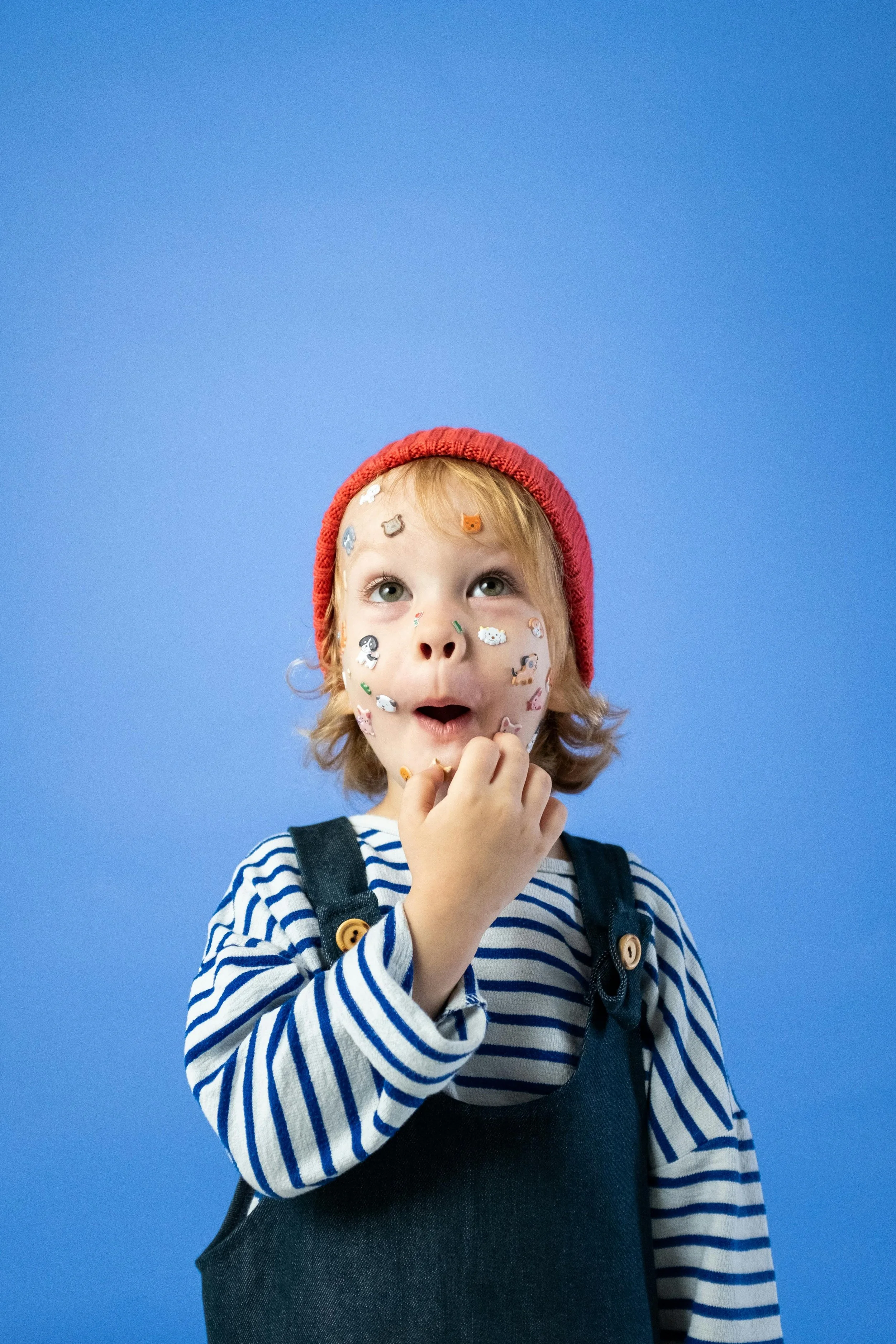 A young child with stickers on face, wearing a red knit hat, striped shirt, and dark overalls, looking upward with a surprised expression against a blue background.