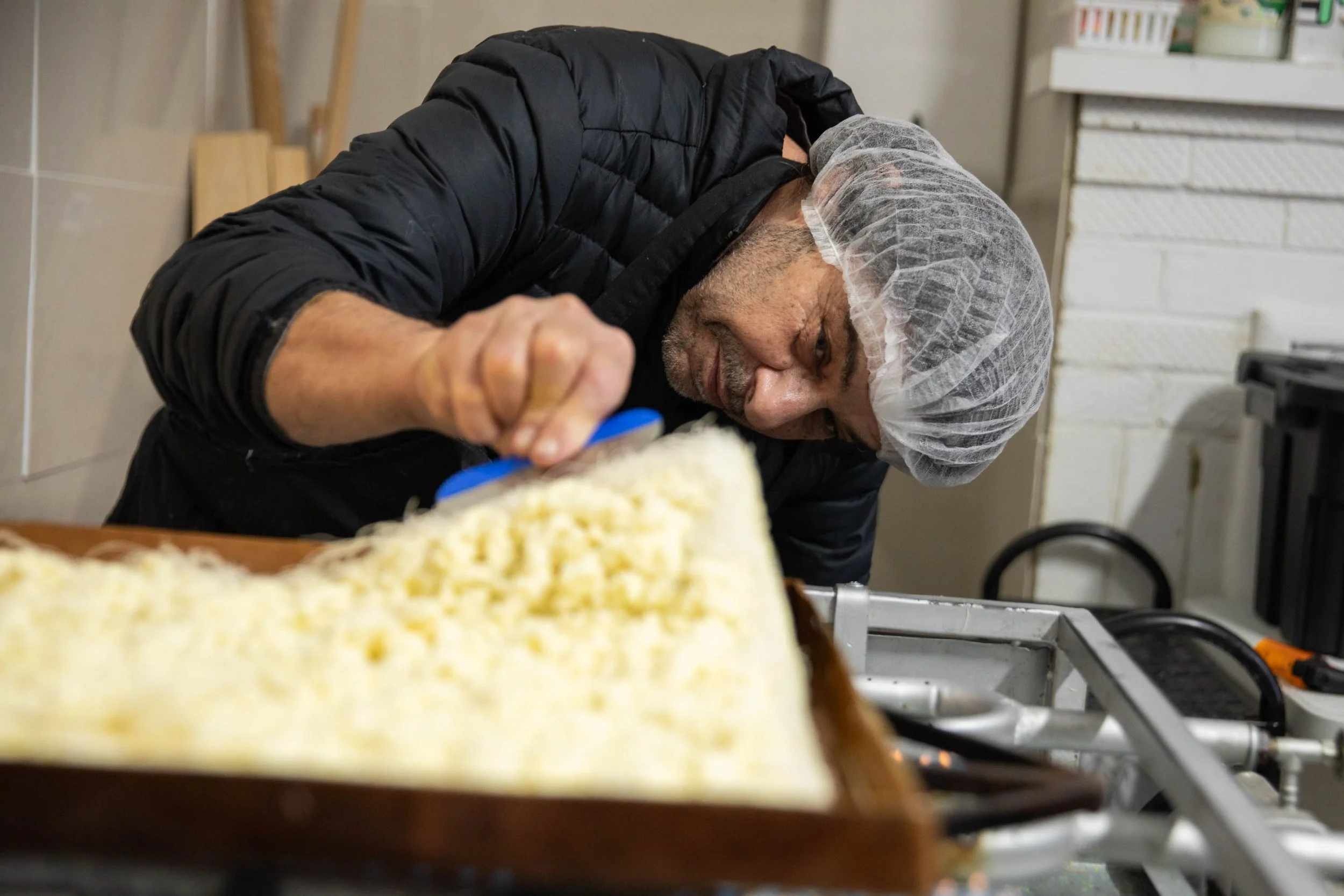 A man wearing a hairnet and black jacket is leaning over a table, working with food. There are trays of food and kitchen storage in the background.