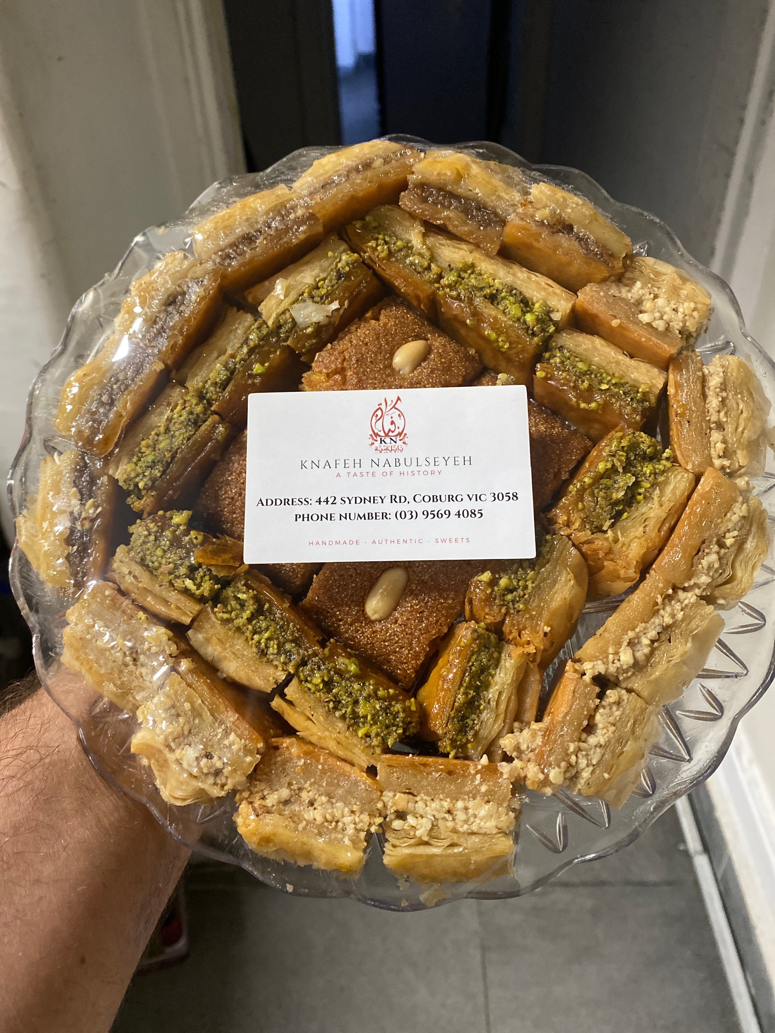 A clear glass platter filled with assorted baklava, topped with crushed pistachios and pine nuts, with a label from Knafeh Nabulseyeh.