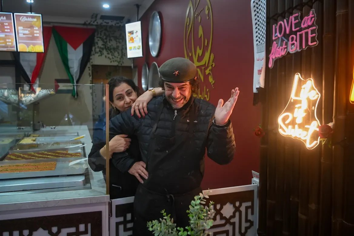 A man in a black jacket and gray beret smiling and waving, with a woman hugging him from behind, inside a restaurant decorated with neon signs that say 'LOVE at First BITE' and 'Life is Sweet' and featuring patterns and flags on the wall.