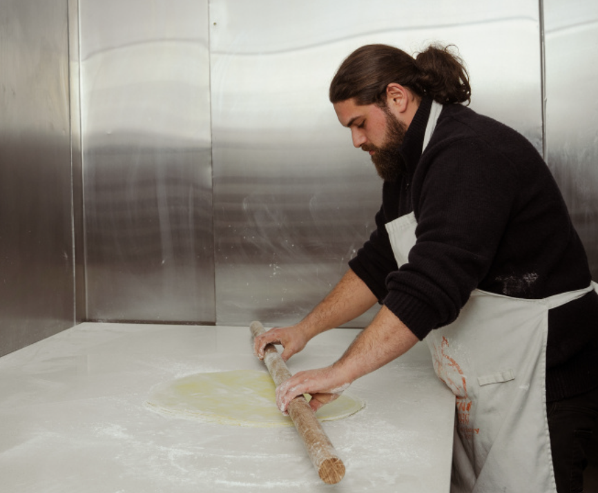 A man with long hair and a beard, wearing a black sweater and an apron, rolling out dough with a rolling pin on a floured surface in a kitchen with stainless steel walls.