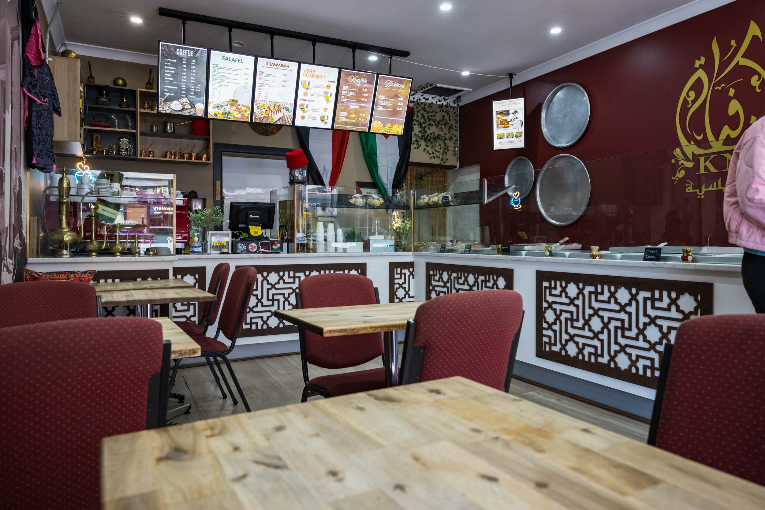 Interior of a restaurant with wooden tables and red upholstered chairs, a counter with display case and decorative elements, and digital menu boards hanging above the counter, with a red wall featuring gold Arabic calligraphy.
