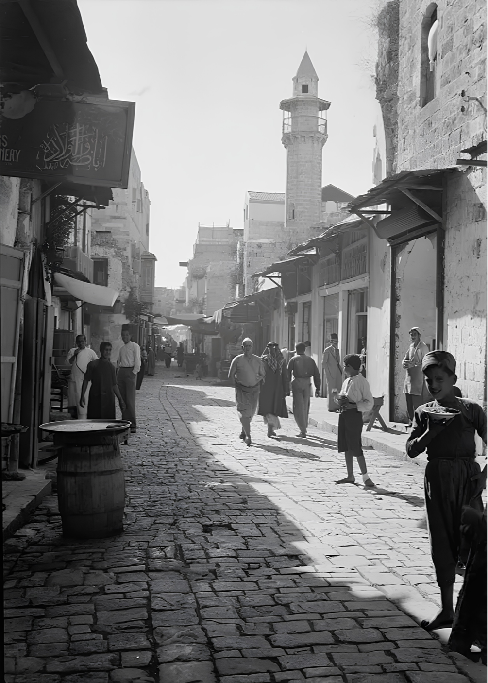 A bustling cobblestone street scene with people walking and talking, old buildings lining the street, and a minaret in the background.
