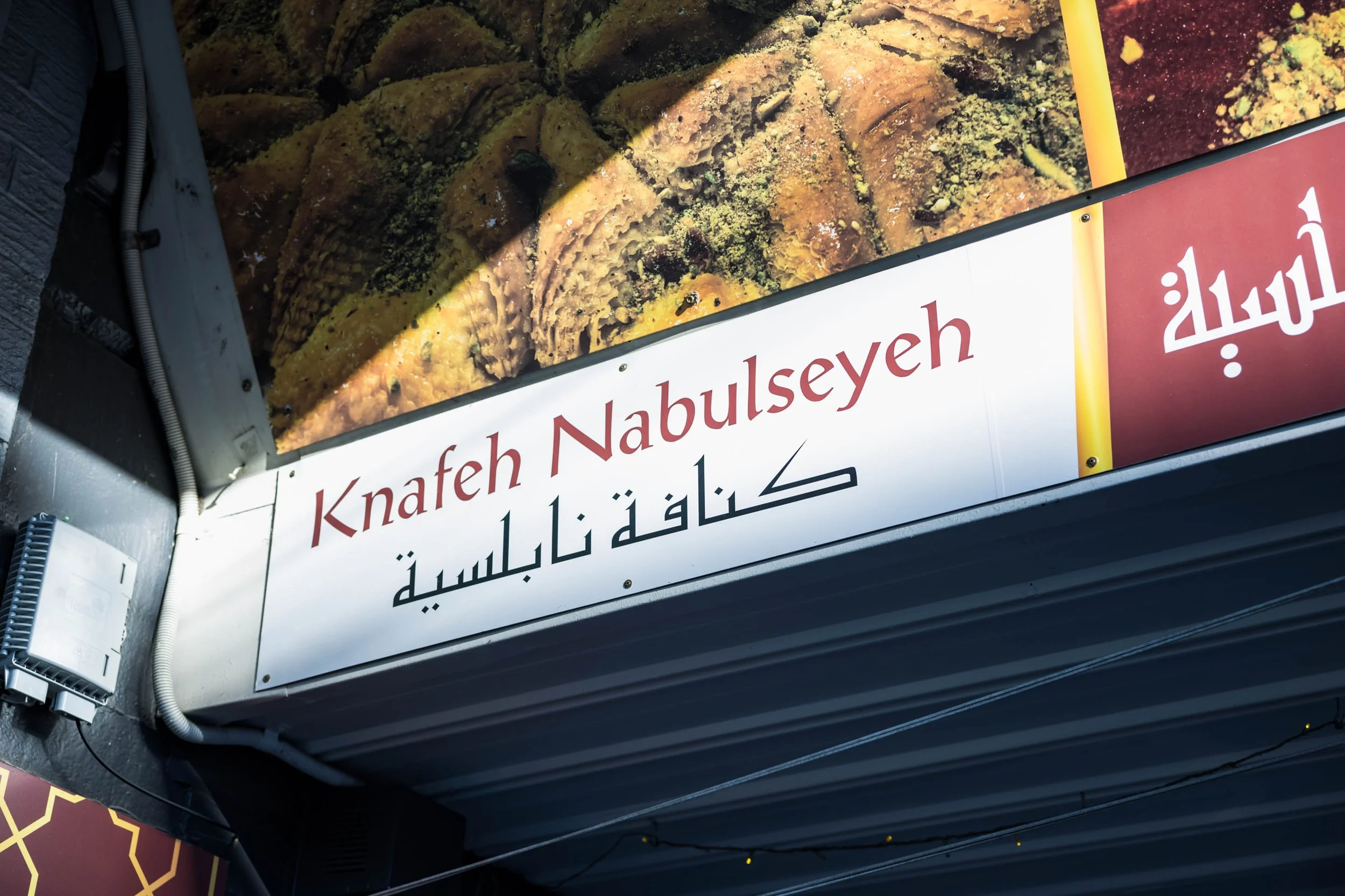 Close-up of a sign with Arabic, Persian, and Latin text, featuring a photograph of traditional baklava desserts coated with honey and sprinkled with crushed pistachios.