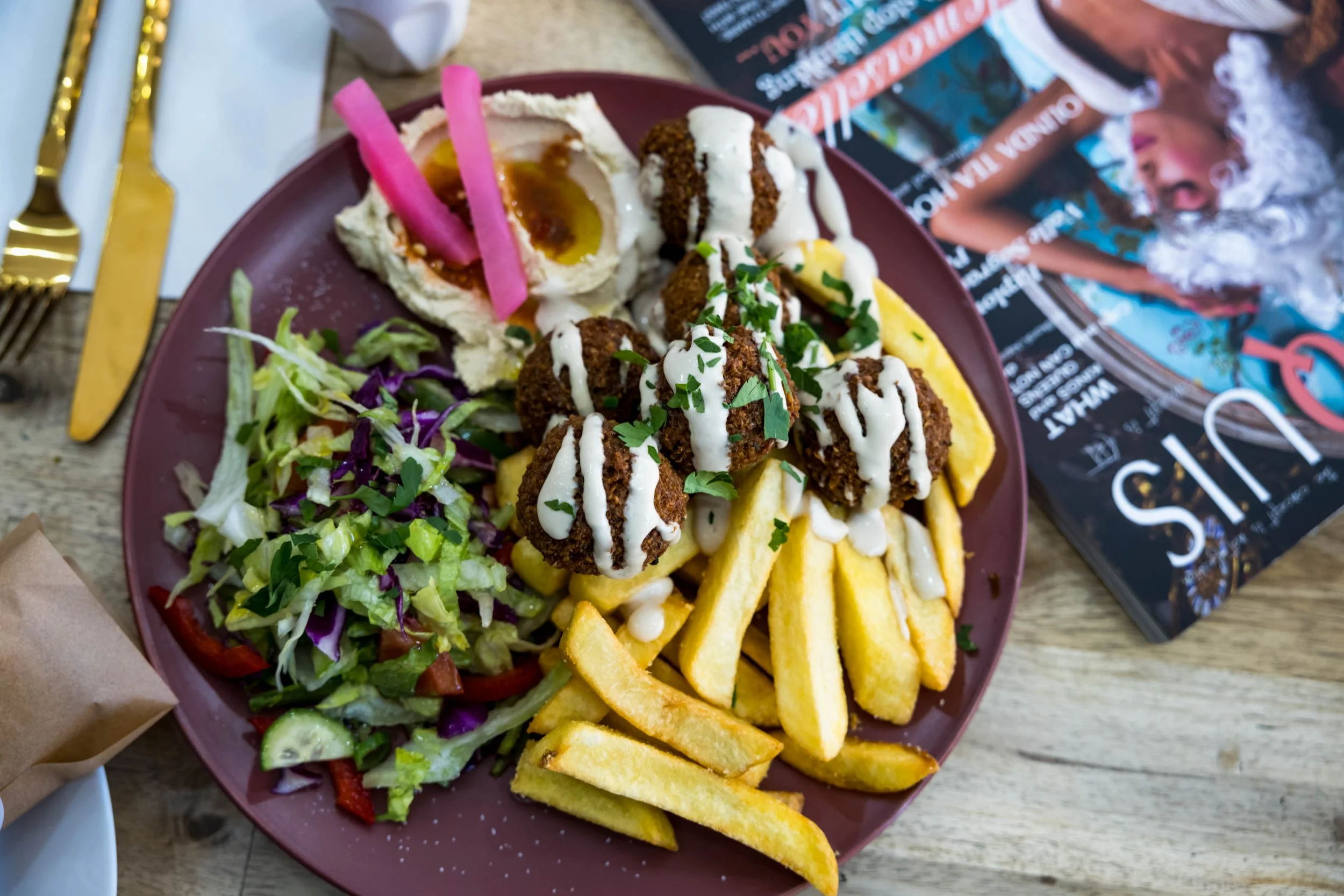 A plate of food with mixed salad, French fries, falafel topped with white sauce, a baked potato with toppings, and pink pickled onions, placed on a wooden table alongside a gold fork and knife, a magazine, and a brown paper takeout bag.