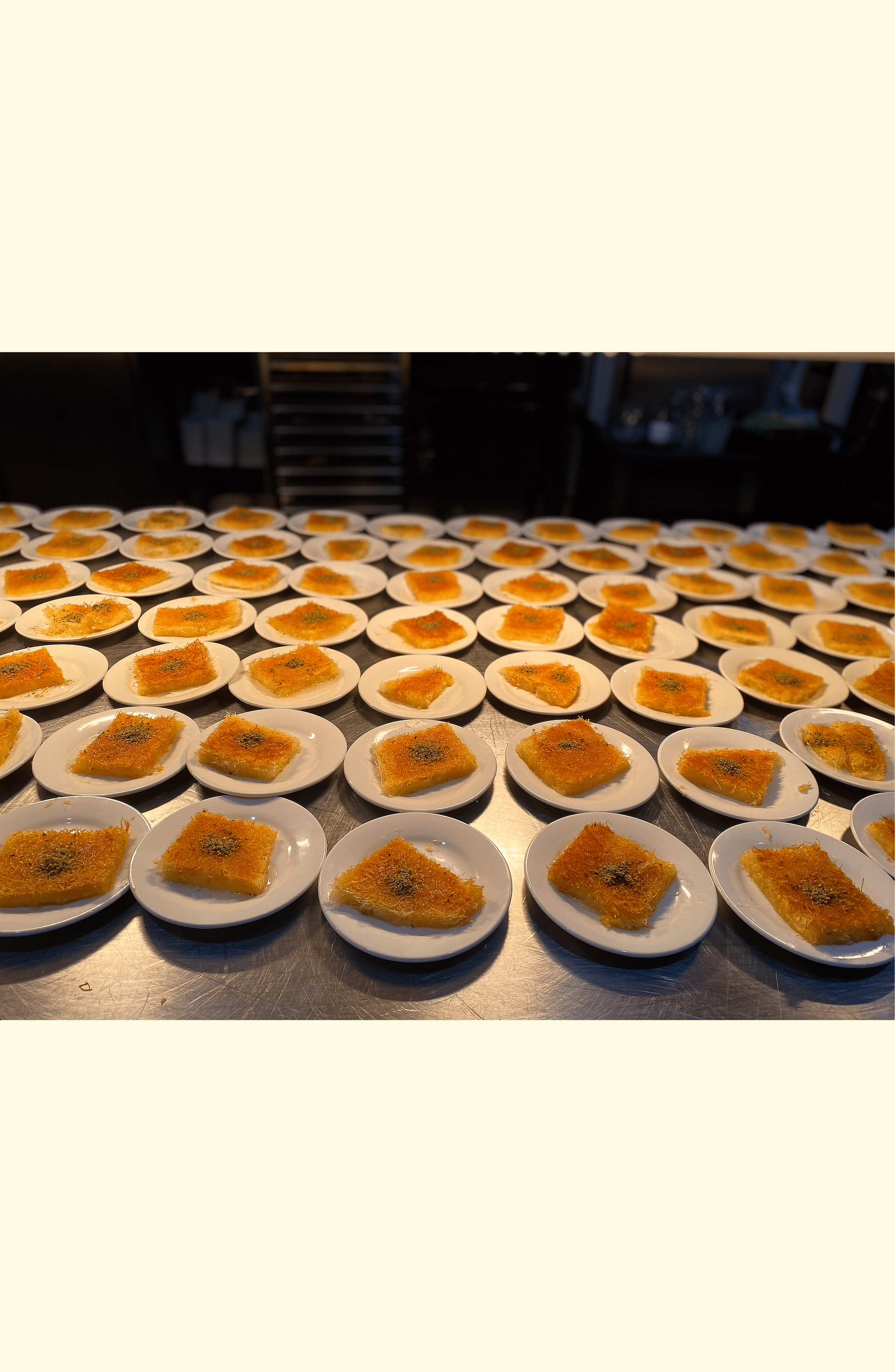 Rows of small white plates with servings of a golden, crispy dessert, garnished with green herbs, on a metal countertop in a kitchen or restaurant setting.