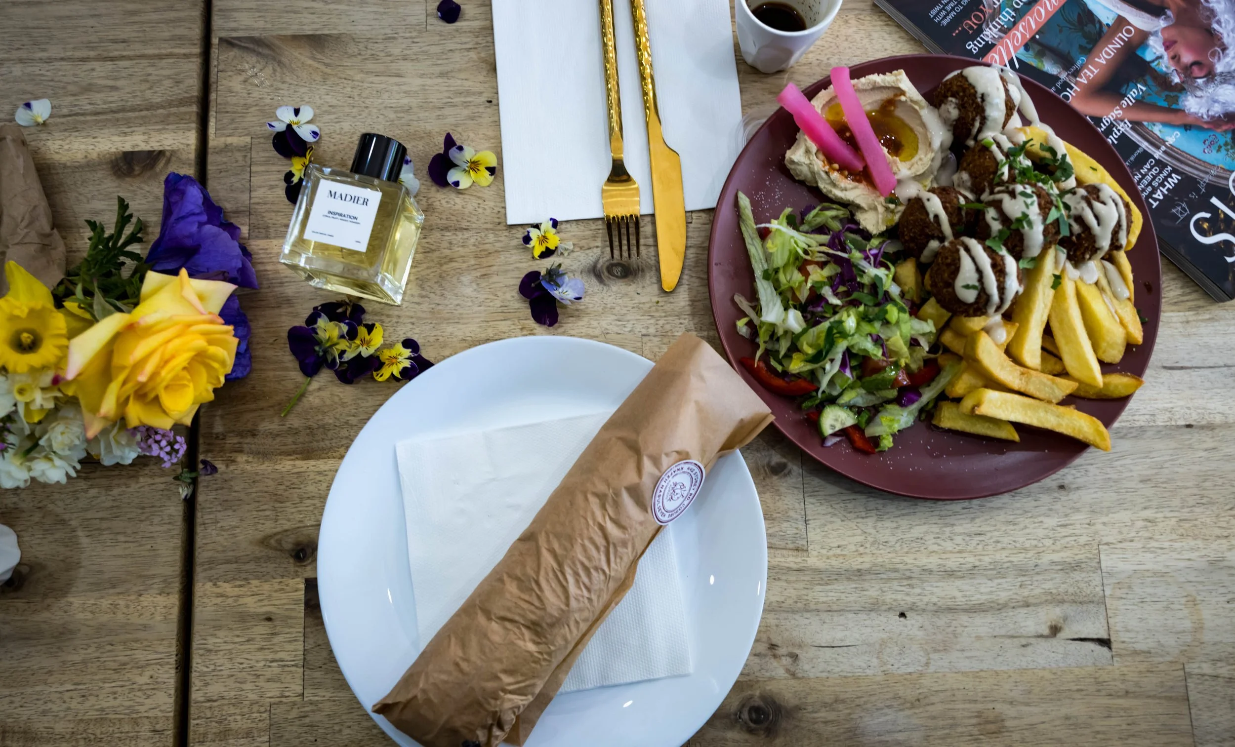 A table setting with a large plate of falafel, fries, and salad, a small cup of coffee, a magazine, a fork and knife with gold handles, a white napkin, a bottle of perfume, and a bouquet of mixed yellow, purple, and white flowers on a wooden table.