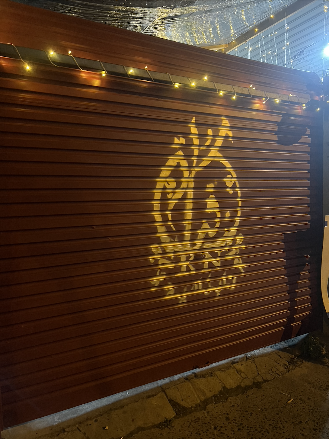 A red metal rolling door with a yellow light projection of a logo or badge. String lights are hanging above the door, and part of a brick sidewalk is visible at the bottom.