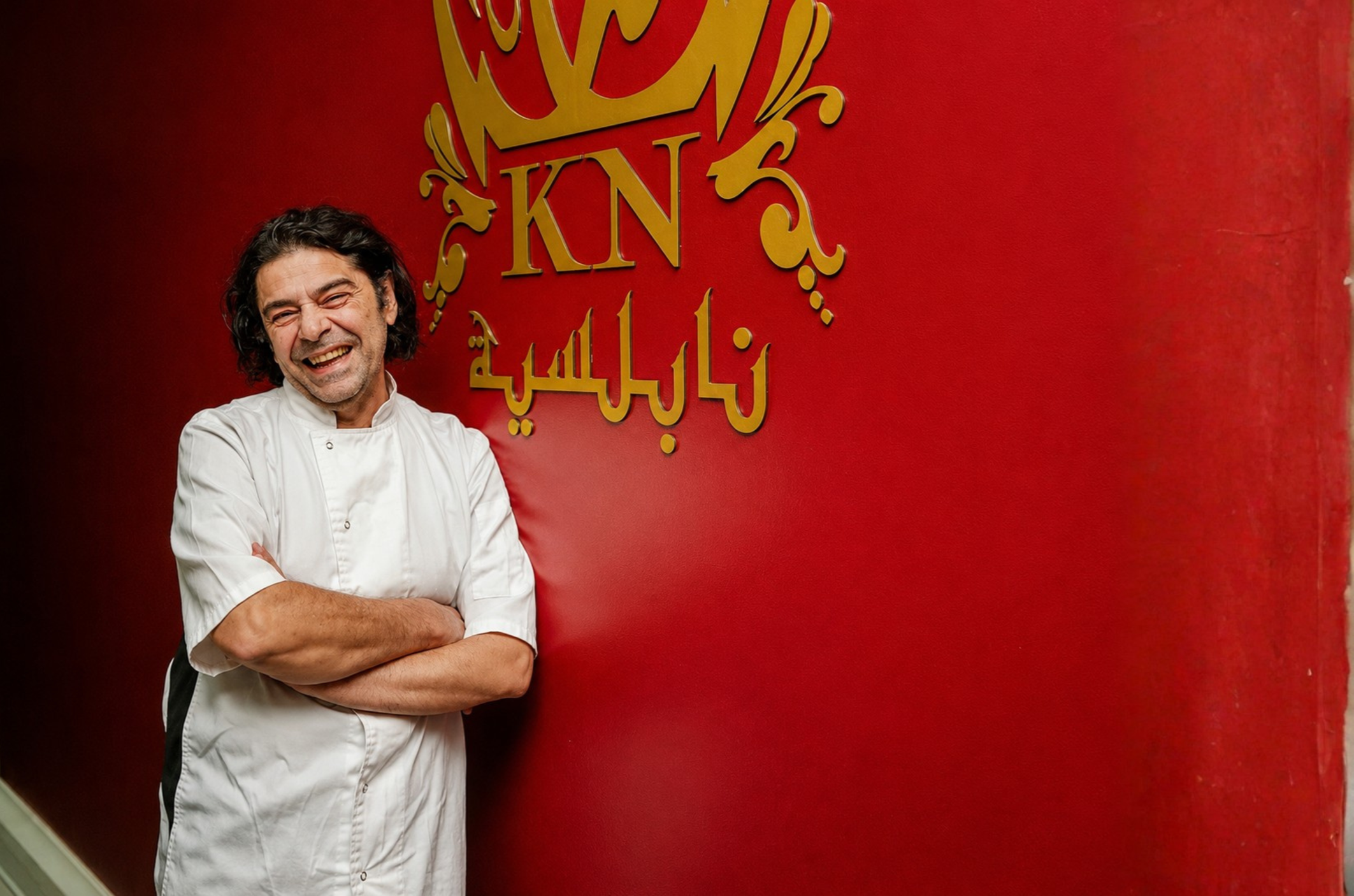 A happy male chef with dark wavy hair, wearing a white chef's coat, standing with arms crossed against a red background with gold lettering.