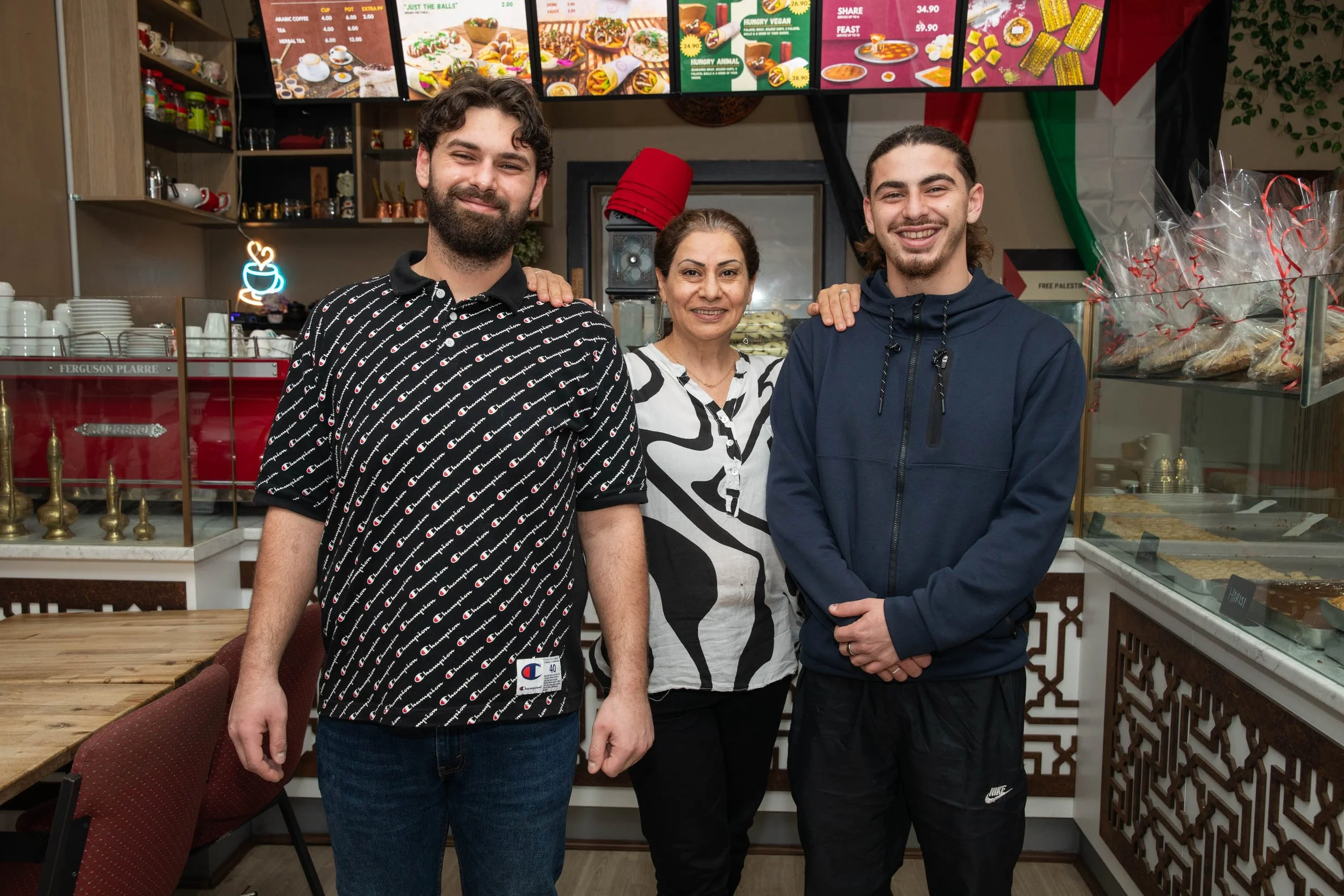 Three people, two young men and an older woman, standing inside a restaurant or cafe, smiling at the camera. The background features menu boards with food images and prices, shelves with cups and decorative items, and bakery items in clear display cases.