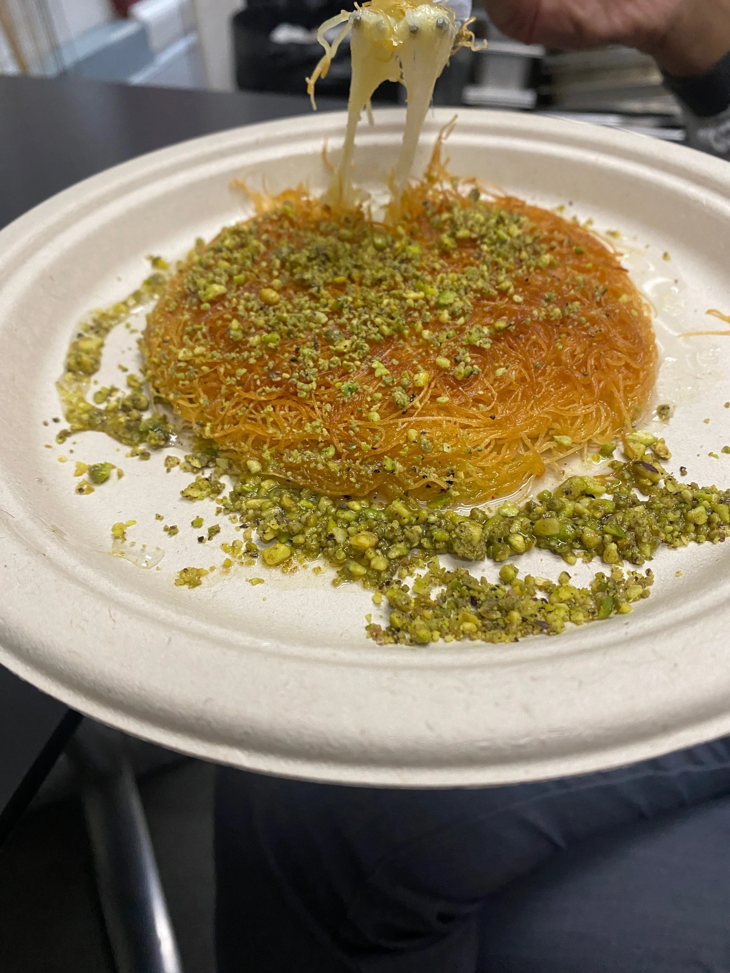 Close-up of a dessert with orange shredded strands topped with chopped pistachios on a white disposable plate.