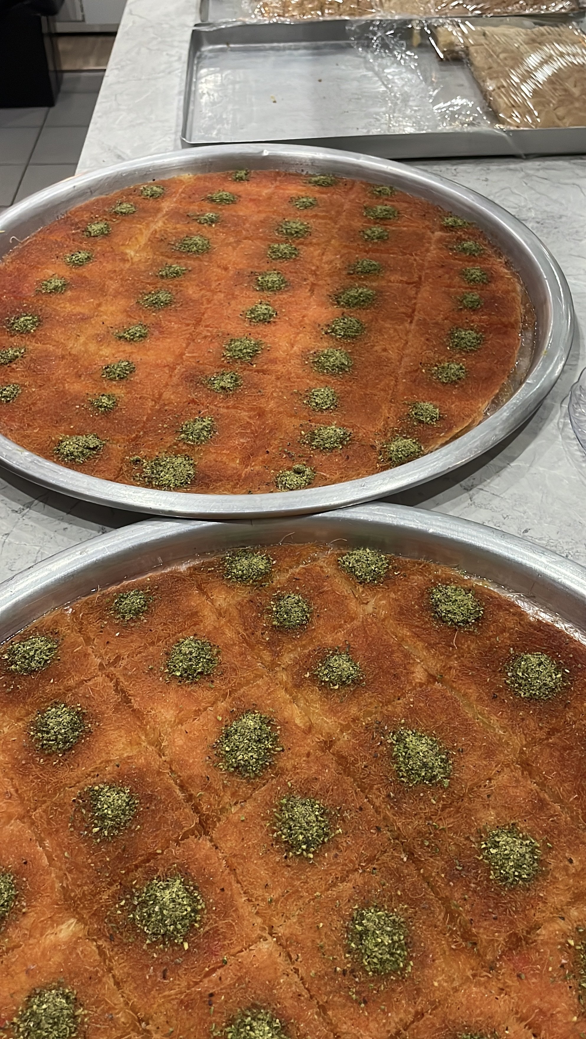 Two large aluminum trays filled with a baked dish topped with green herbs, possibly parsley or cilantro, and cut into slices. The trays are on a marble countertop with a tray of baked goods or cookies in the background.