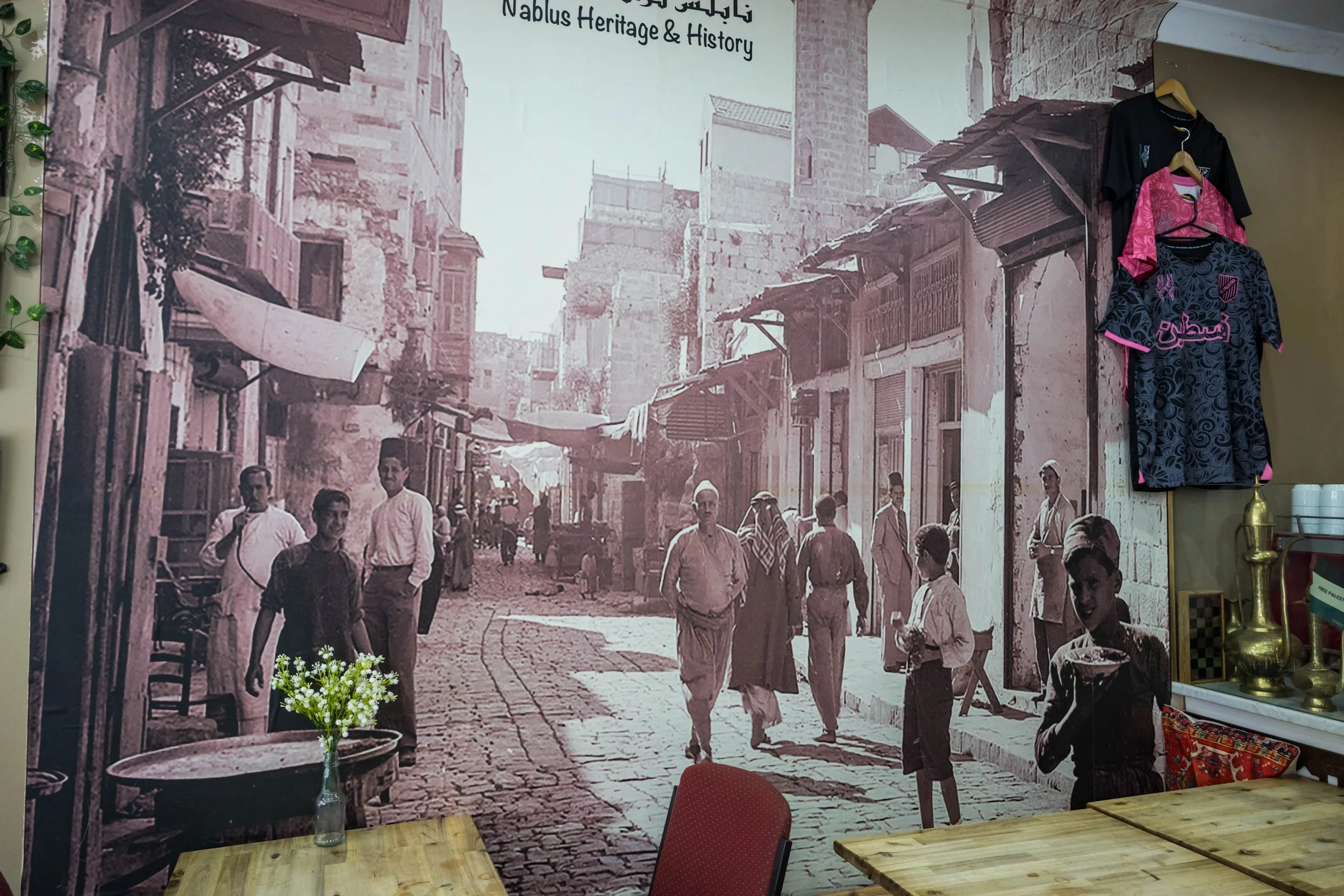 A large black and white historical photograph of a busy street scene with people walking and buildings showing signs of wear, located on a large wall in a room. There are some clothes hanging on the right side, and a small table with a red chair and a flower in the foreground.