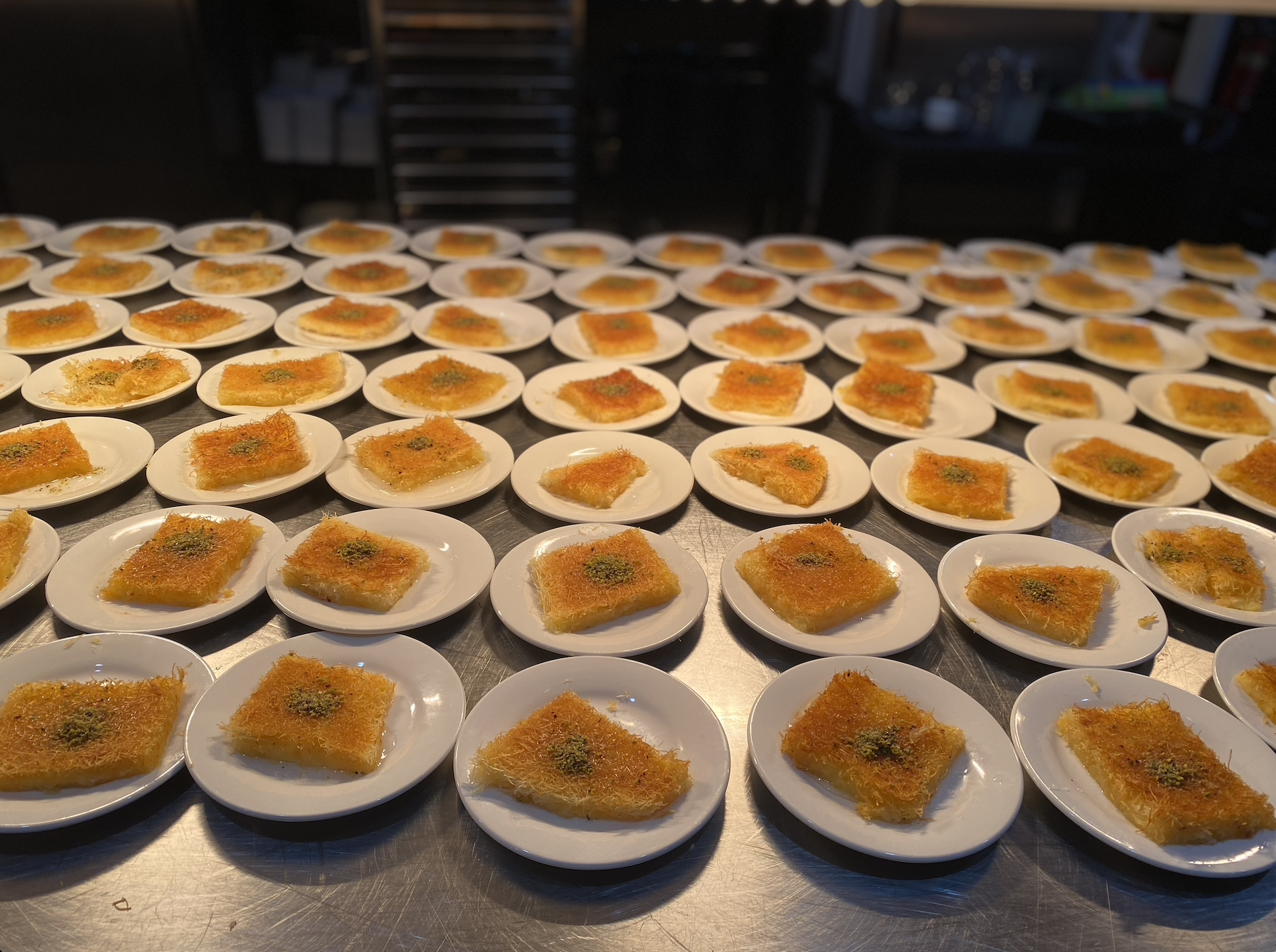Multiple small white plates with pieces of a golden crispy dessert topped with green garnish, arranged on a stainless steel table. The background appears to be a kitchen environment.