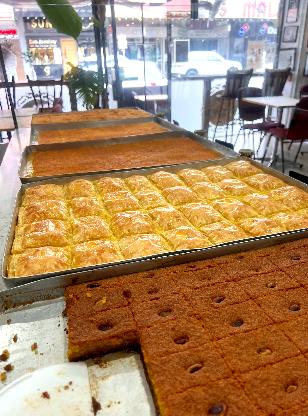 Various trays of baked desserts including baklava, pink layered pastry, and chocolate chip squares inside a bakery or cafe, with tables, chairs, and street view through large window in the background.